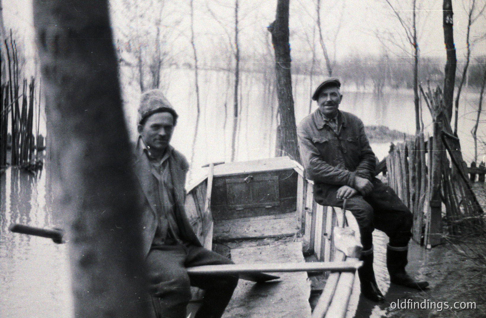 Two men seated in a small, makeshift boat in a flooded landscape. One wears a beret, the other a wool cap. Heavy boots suggest damp conditions. Sparse trees and a distant shoreline define the scene. Likely early-mid 20th century. A visual record of adapting to environmental conditions.