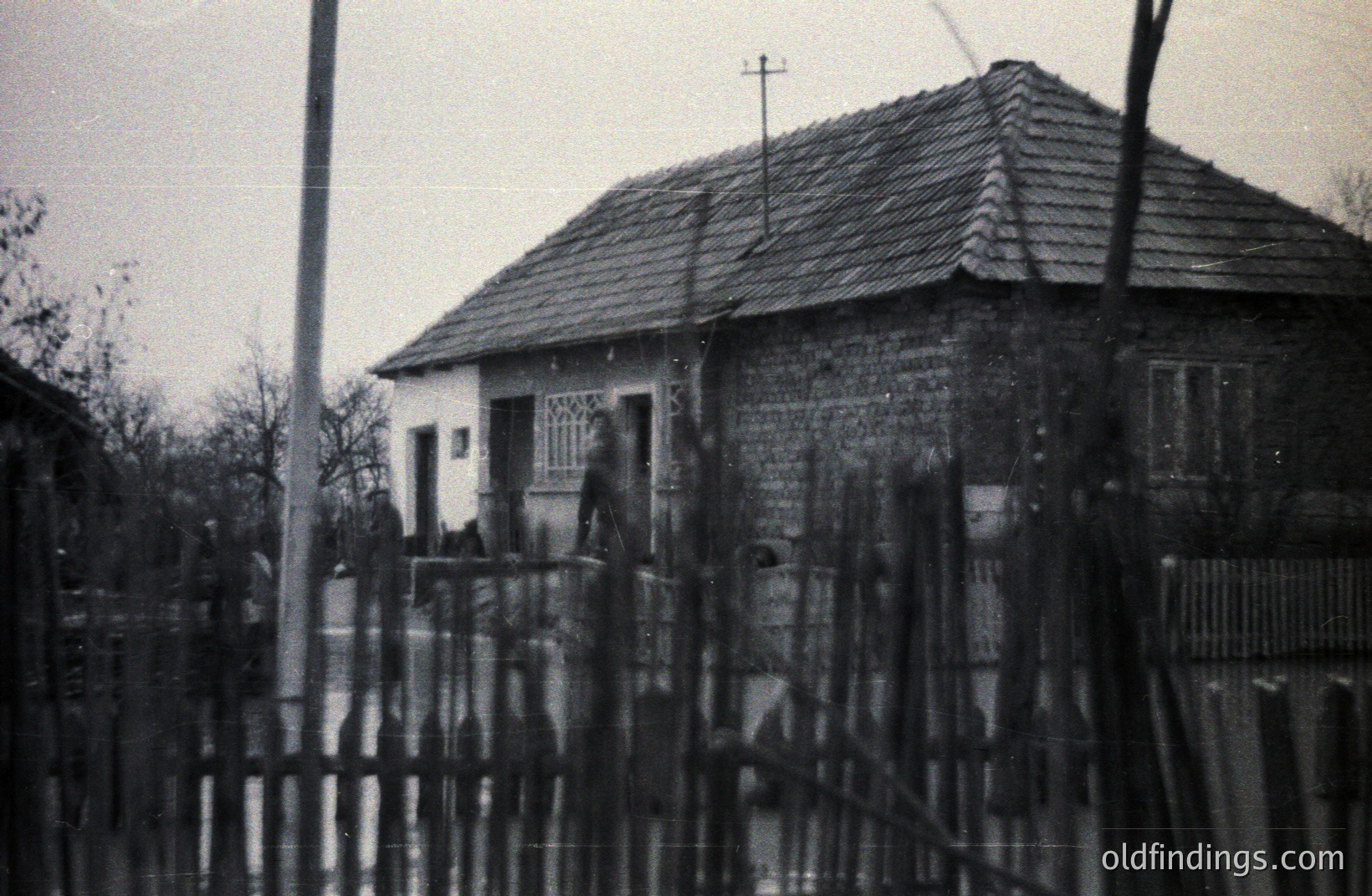 A modest, single-story house with a steeply pitched, shingled roof is framed by a weathered wooden fence. Utility pole and bare trees are visible. Likely rural Eastern Europe, possibly 1950s-1970s, judging by architectural style and photographic technique. Demonstrates vernacular architecture.