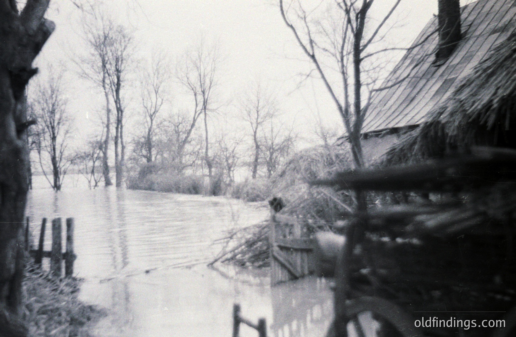 Flooded landscape view with a weathered wooden structure partially visible in the foreground. Bare trees line the water's edge; likely post-winter. The scene evokes a rural, possibly agricultural context. Grainy texture suggests an older, possibly amateur photograph.