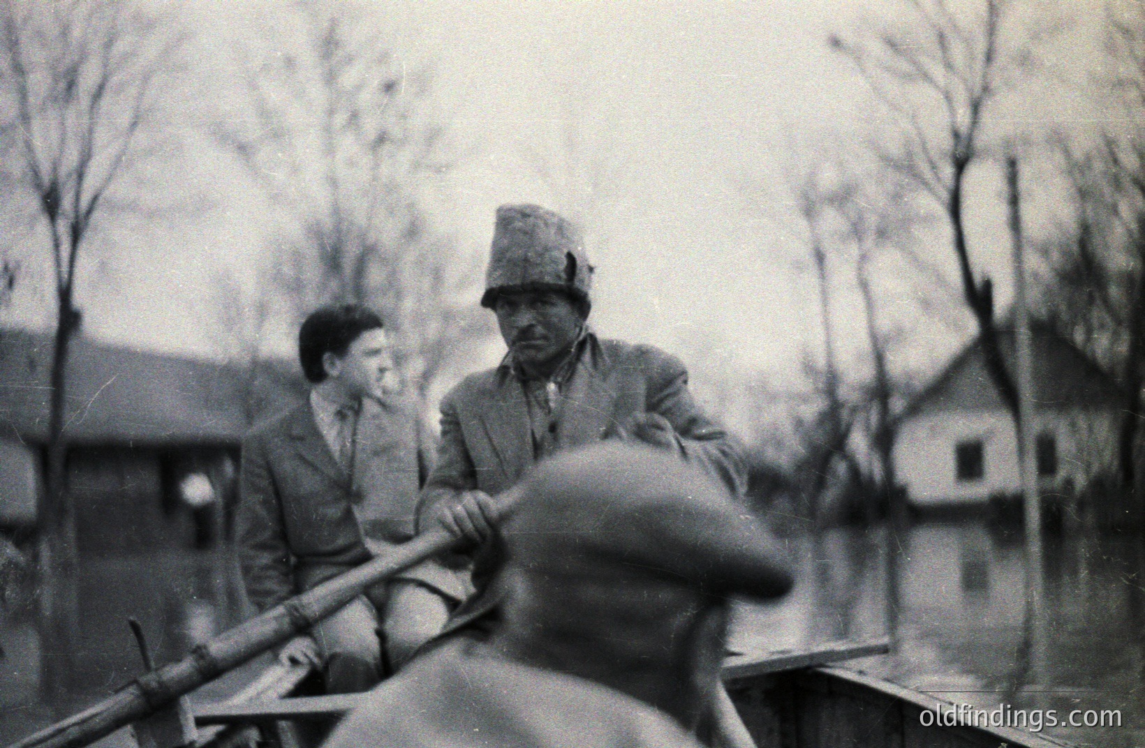 A man rows a small boat carrying a young boy through floodwaters, likely post-war period. Sparse trees and a simple building are visible in the background. The grainy image suggests a documentary or personal snapshot. The style is reminiscent of mid-20th century rural photography.