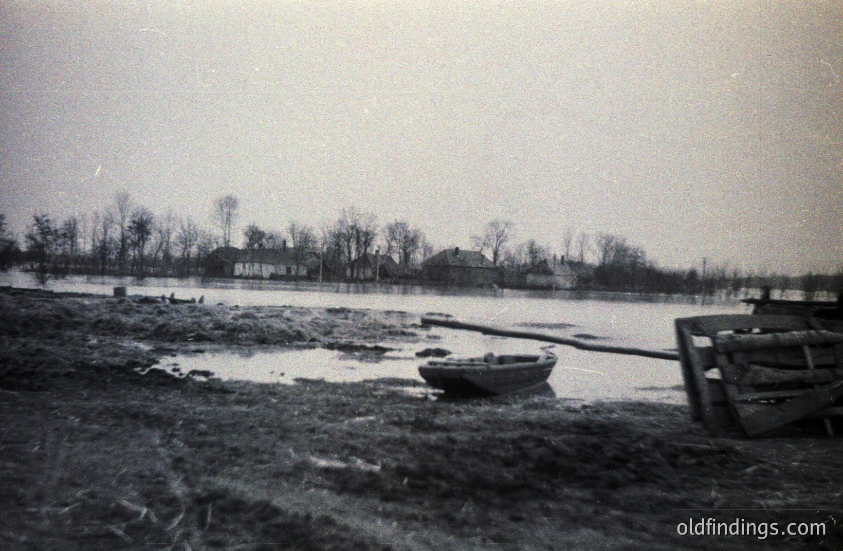 Flooded landscape with a small rowboat beached on muddy ground. Several simple, wood-frame houses are visible in the distance, partially submerged. Likely a rural scene, possibly post-flood event. Appears to be a candid, documentary-style image. Estimated 1950s-1970s.
