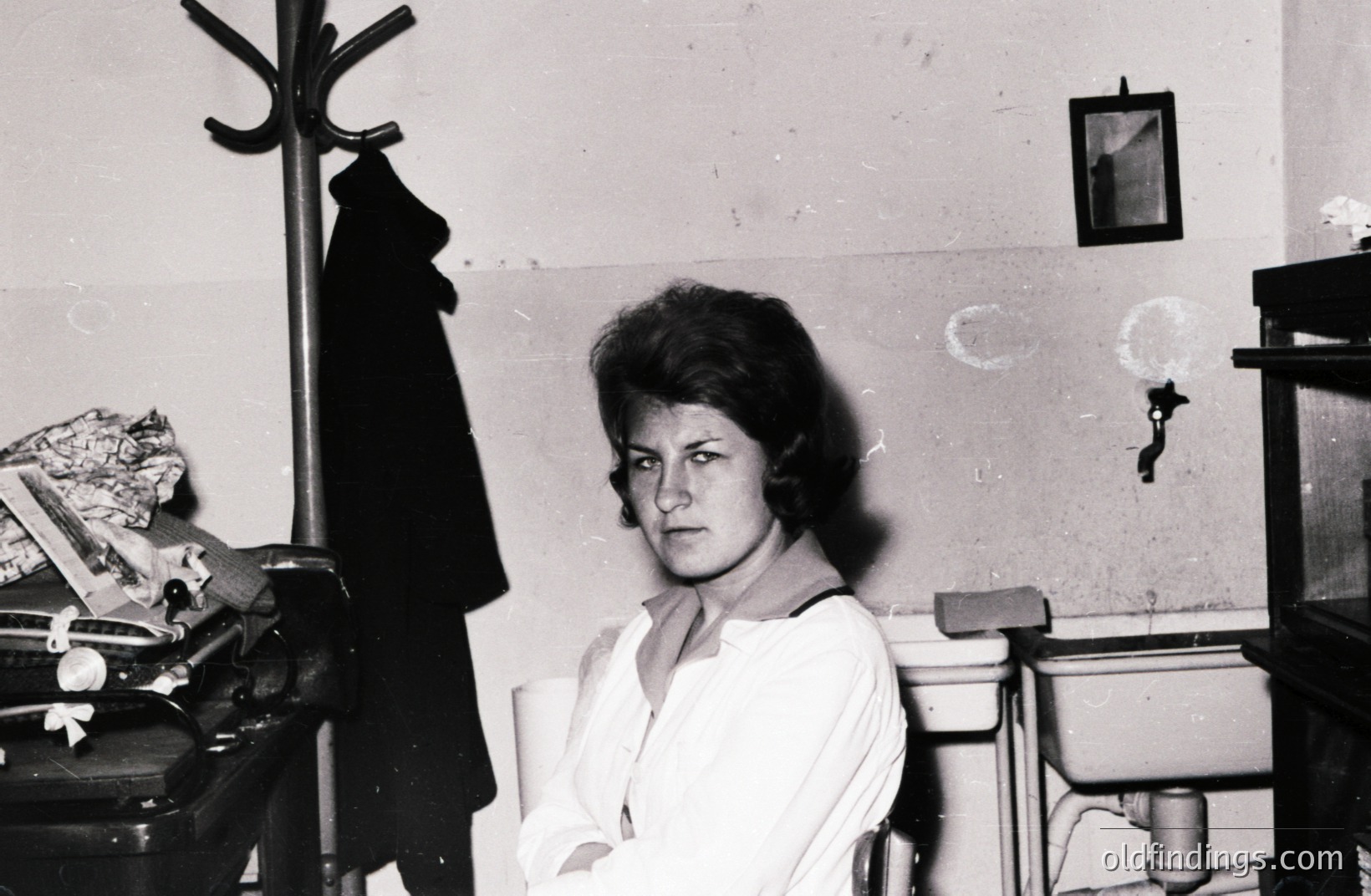 A young woman sits in what appears to be a small, utilitarian room, possibly a caretaker's office. She wears a crisp white blouse and has a neat, short hairstyle typical of the 1960s. A coat hangs on a nearby hat rack; a small framed photo is on the wall. Simple, stark setting.