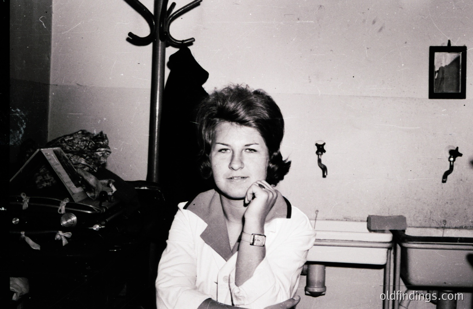 A young woman with a short, styled haircut sits pensively, chin resting on her hand, in what appears to be a hallway or entryway. She wears a white, collared shirt and a wristwatch. Visible details: coat rack, wall hooks, a framed picture, and a glimpse of a decorative object on a nearby shelf. Likely 1960s.