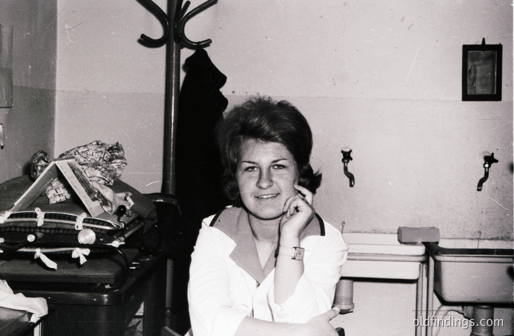 A young woman with a bouffant hairstyle is posed for a portrait, arms crossed, in what appears to be a public restroom or changing area. Vintage luggage and a coat rack are visible. Likely 1960s, evoking a mid-century travel aesthetic.