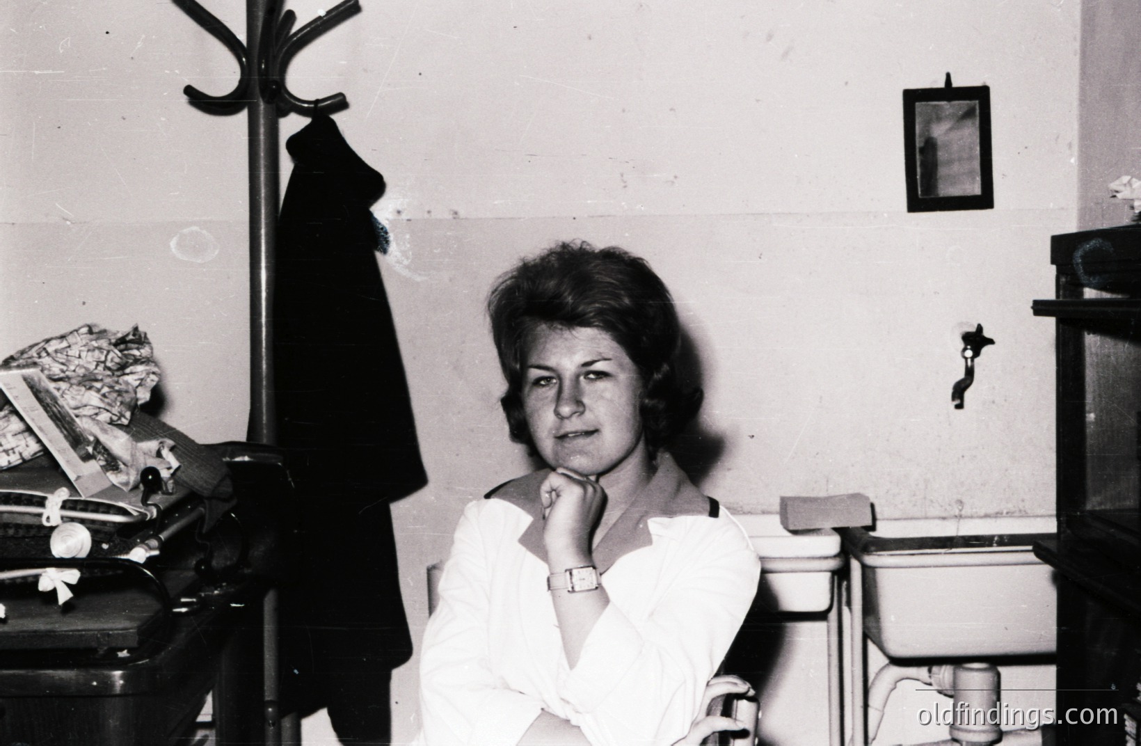 Black and white portrait of a young woman with a short, textured hairstyle, arms crossed, posed in what appears to be a dressing room or utility space. A coatrack with a dark coat hangs in the background. Visible elements suggest a mid-century domestic interior.