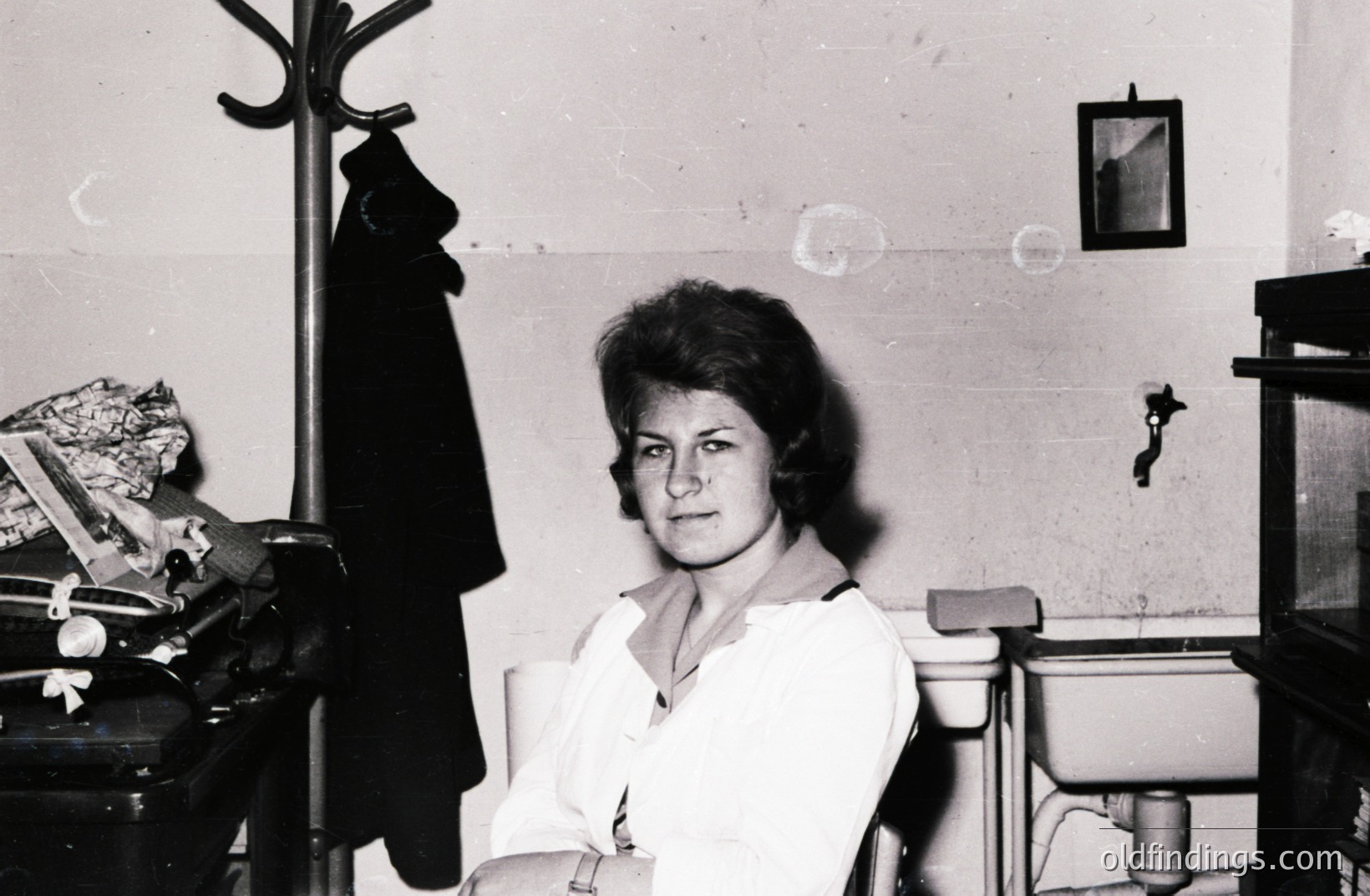 A woman sits on a utility sink cover, arms crossed, in what appears to be a service area. She wears a white collared shirt. A coat rack and a cabinet are visible. Minimalist decor, possible institutional setting. Likely 1960s or 1970s. Good reference for period design.