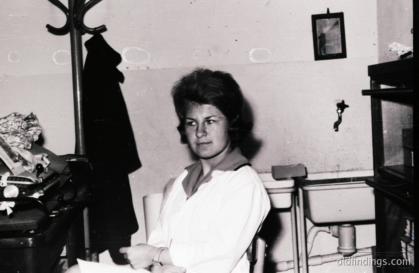 A young woman in a crisp white uniform sits in a simple chair, posed near a washbasin and shelving unit. The photograph, likely taken in the 1960s or 70s, shows a functional interior, suggesting a medical or institutional setting. A coat hangs on a nearby rack.