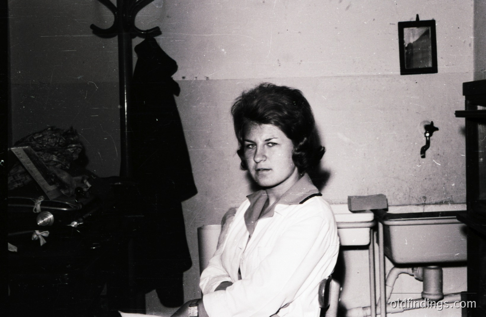 A young woman in a white shirt and dark tie sits facing the camera in a stark, institutional setting. A coat rack with a dark coat, and a sink with a small mirror are visible. Likely a medical or educational facility, 1960s. Photo displays age-related deterioration.