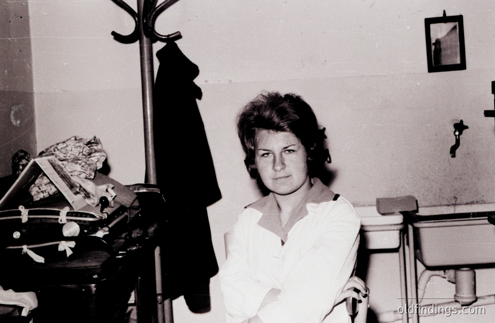 A portrait of a young woman in a simple blouse, posed in what appears to be a school or institutional setting. Note the coat rack and metal desk. Likely 1960s/70s aesthetic, classic mid-century style. A framed picture hangs on the wall.
