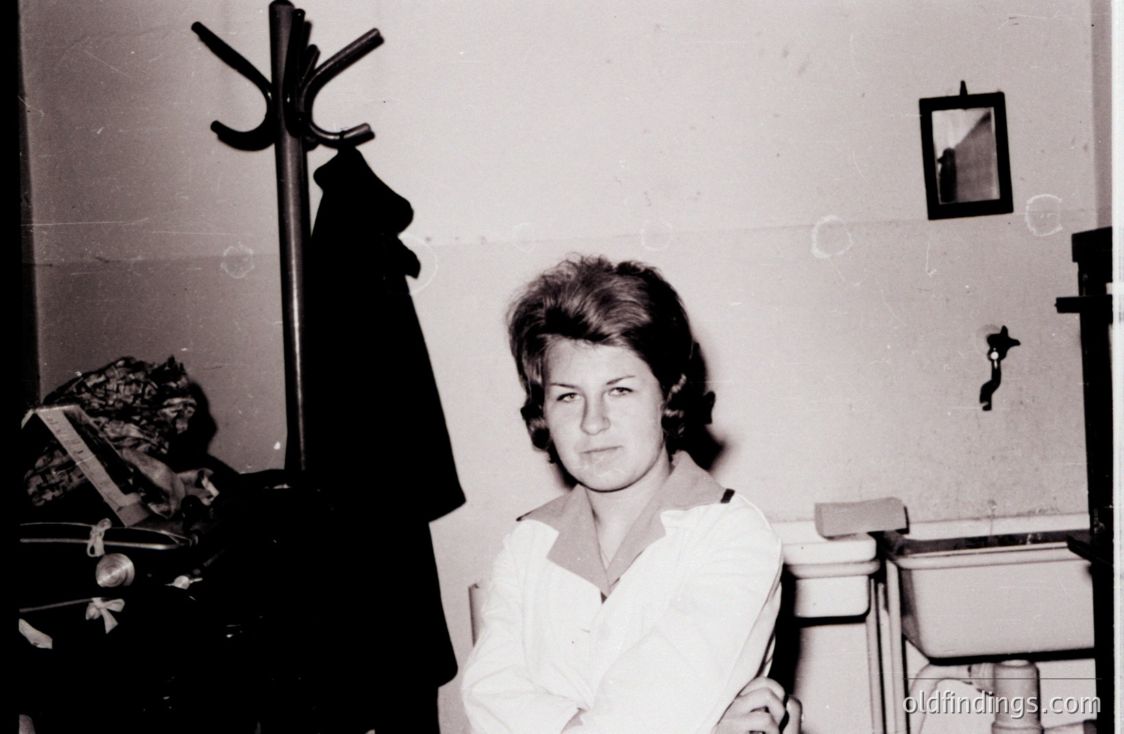 Black and white portrait of a young woman with a short, layered hairstyle, likely 1960s. She wears a light-colored collared shirt. A coat rack and small mirror are visible in the background, suggesting a hallway or entryway. A sense of domesticity.