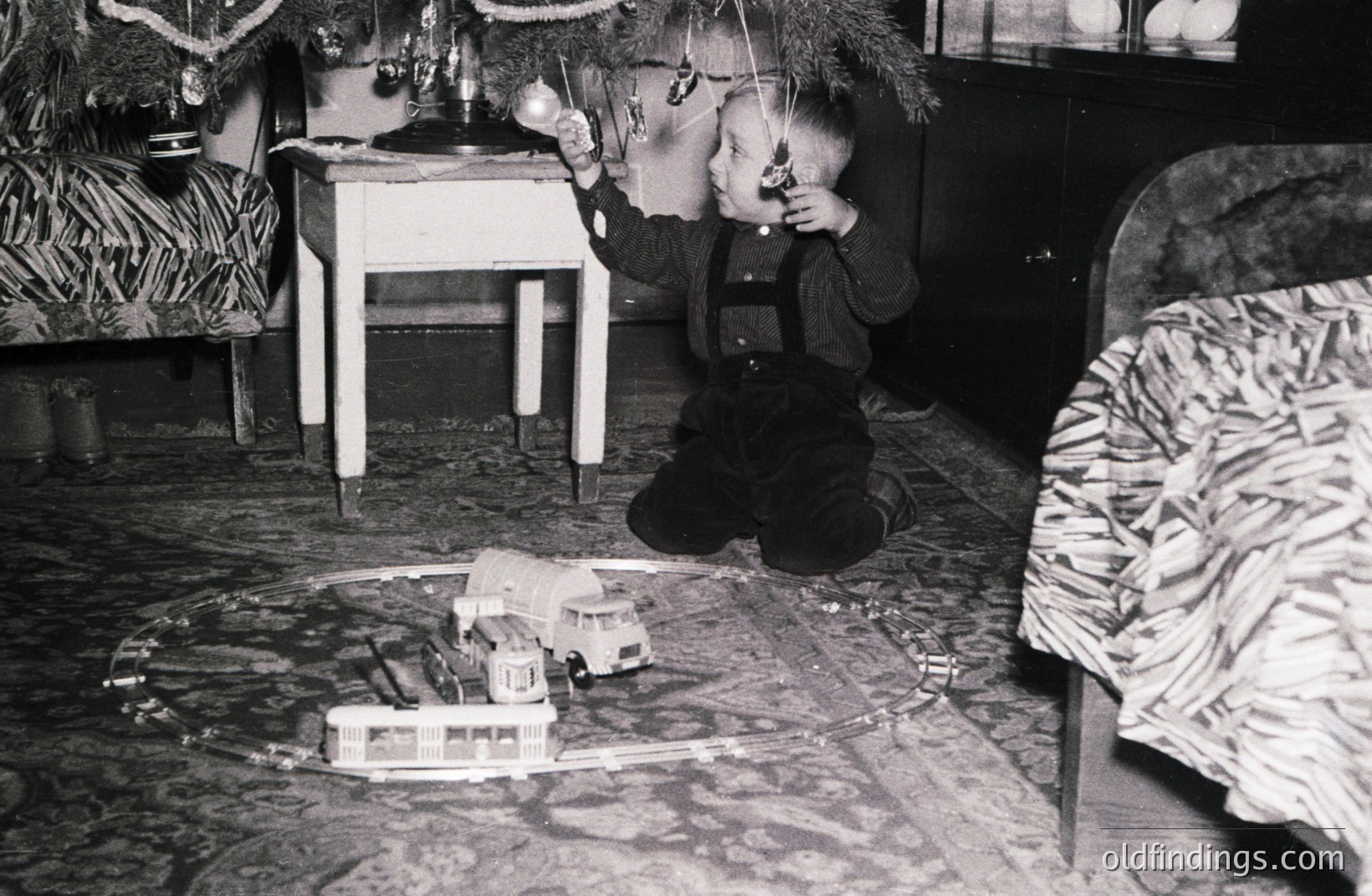 A young boy, dressed in overalls, plays with a vintage model train set beneath a decorated Christmas tree. Details suggest a mid-20th century interior, possibly the 1940s-1960s, with ornate furniture and a cozy atmosphere. Likely family holiday scene.