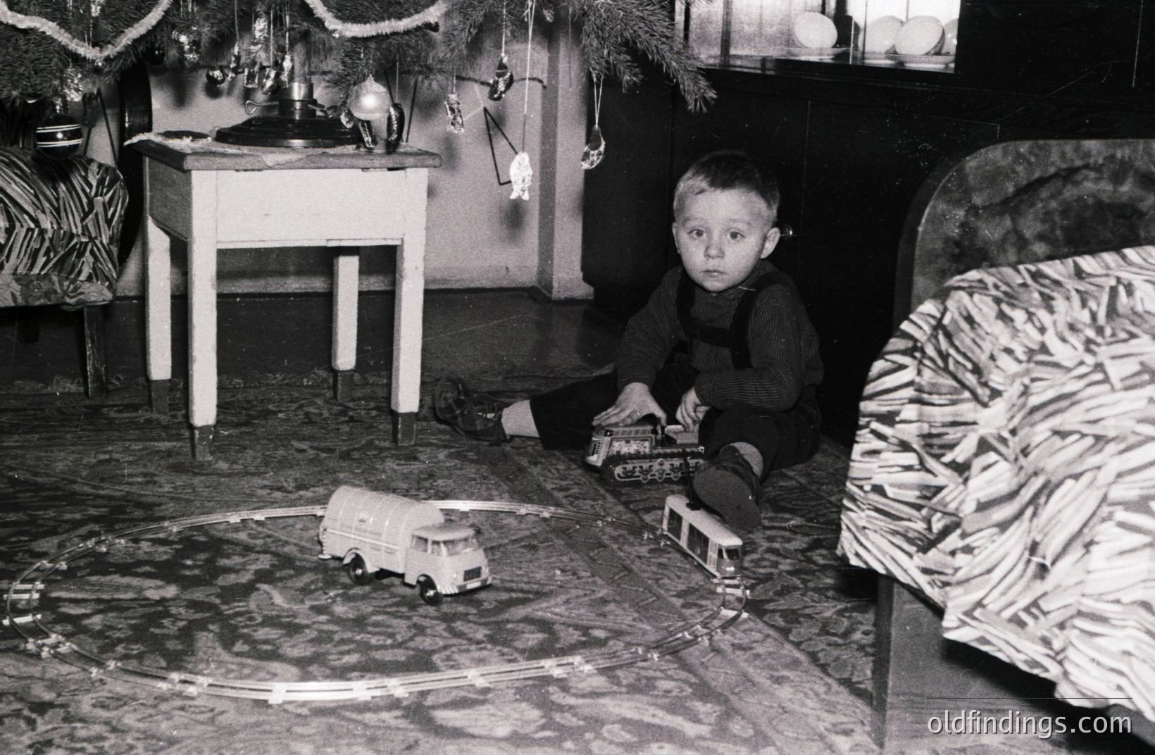 A young boy, wearing overalls, sits cross-legged amidst a miniature train set on an ornate rug. Decorated with sparse ornaments, a Christmas tree stands nearby. The room’s aesthetic suggests a mid-century interior. Likely a family snapshot documenting childhood joy.