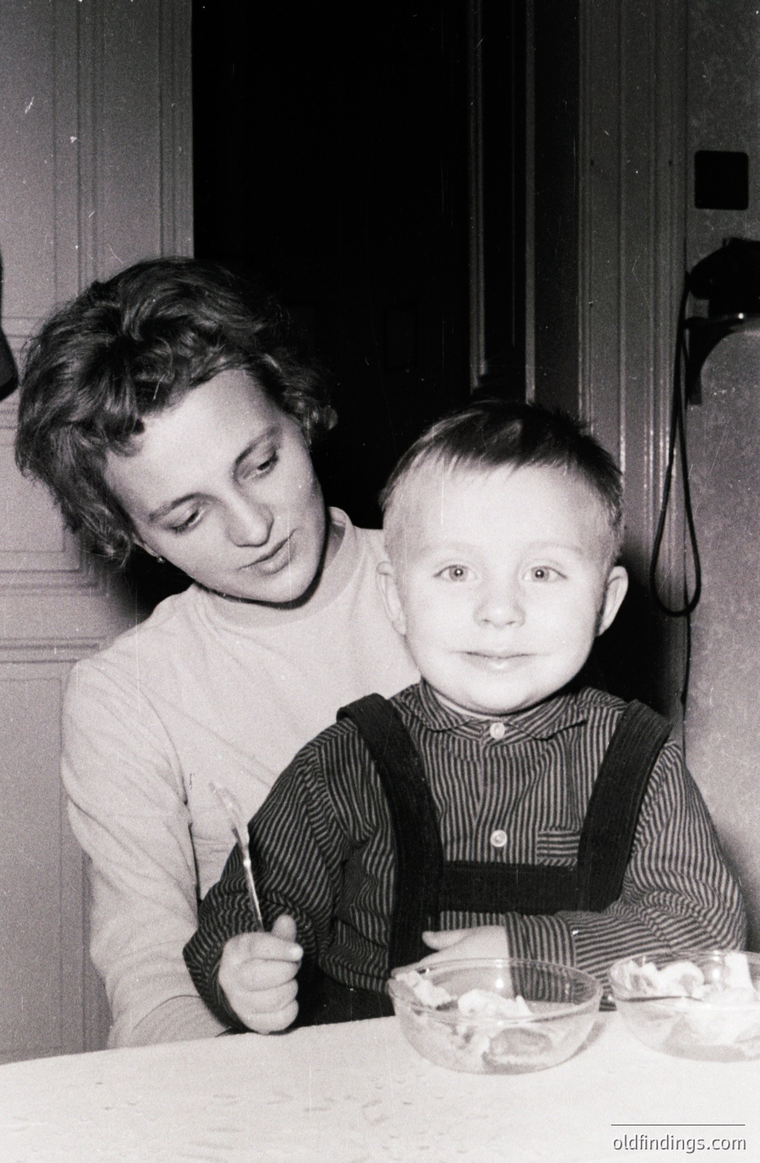 A young woman assists a toddler at a table, both appearing attentive and engaged. The child wears a striped shirt with suspenders and holds a spoon. Two clear bowls with food are visible. Likely a domestic scene, indicative of mid-20th century family life.