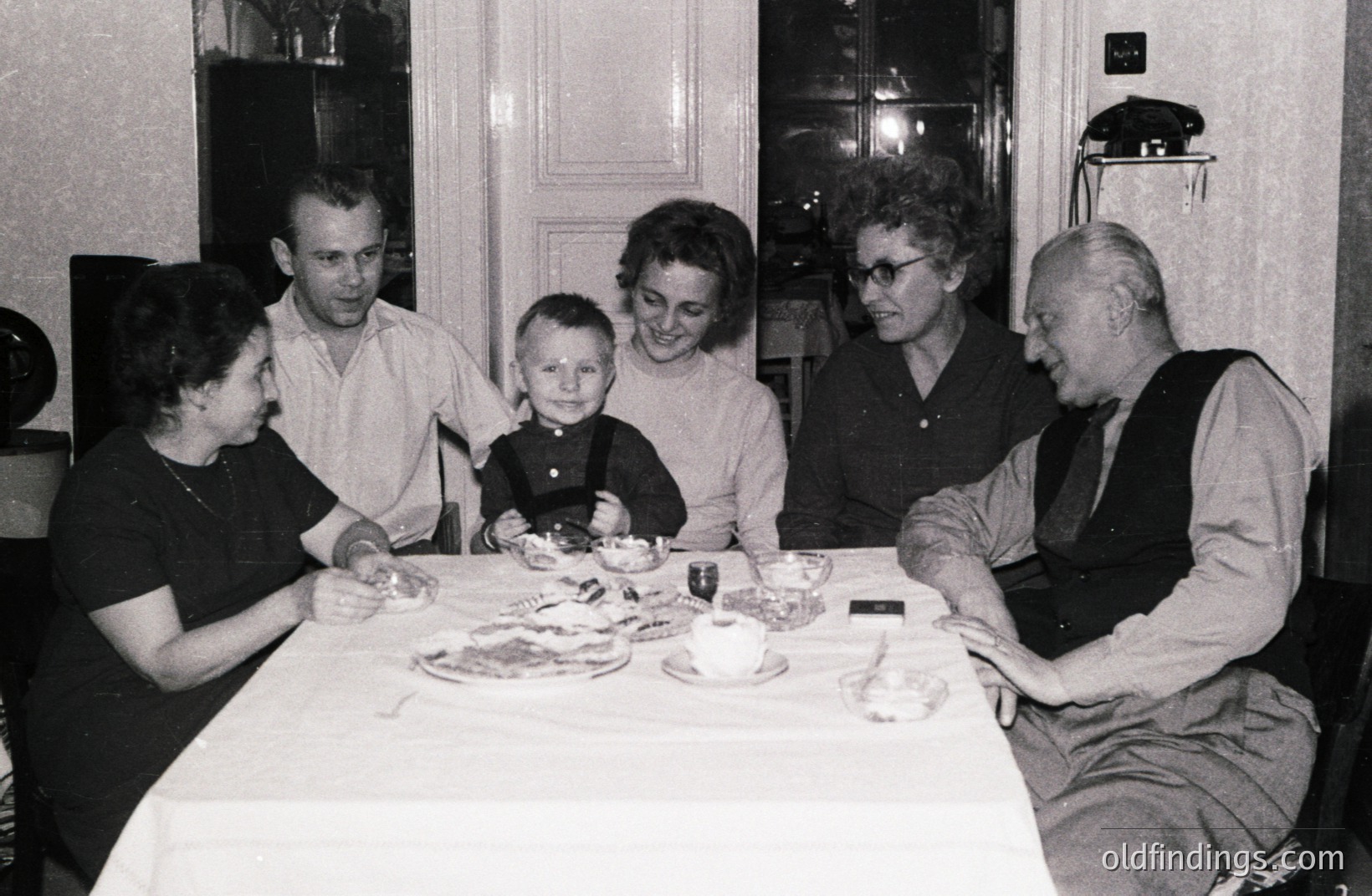 Family gathered around a table laden with food, likely a celebratory meal. Five individuals are visible, including a young boy in overalls. Interior setting with dark wallpaper and a vintage telephone wall mount suggest a mid-century home. Likely 1960s-1970s.