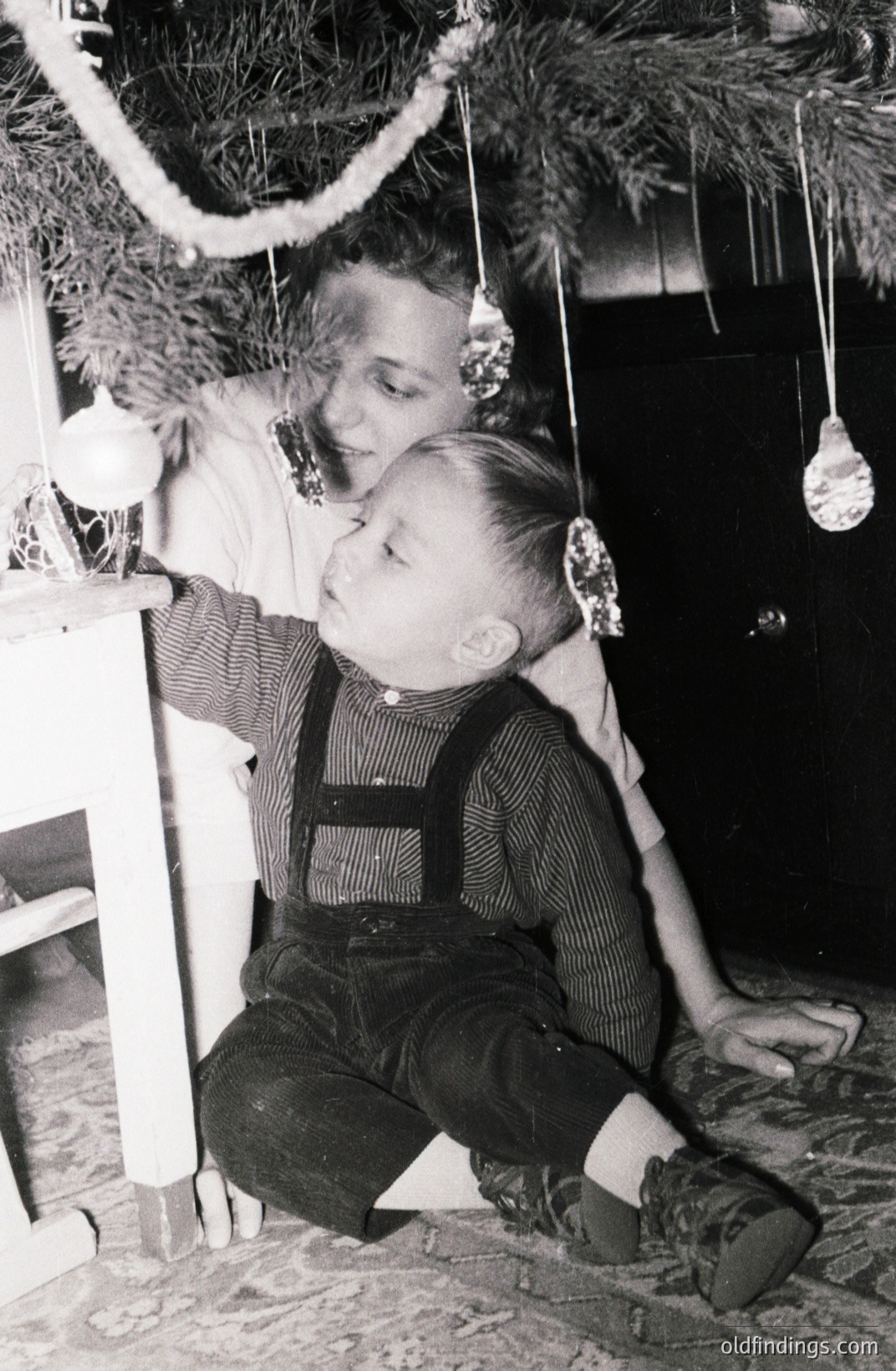A young boy in overalls sits near a decorated Christmas tree, partially obscured by a smiling adult. Simple ornaments hang from the branches. Likely a domestic scene, potentially from the 1940s or 50s. Evokes nostalgia & family tradition. A useful reference for design or historical research.