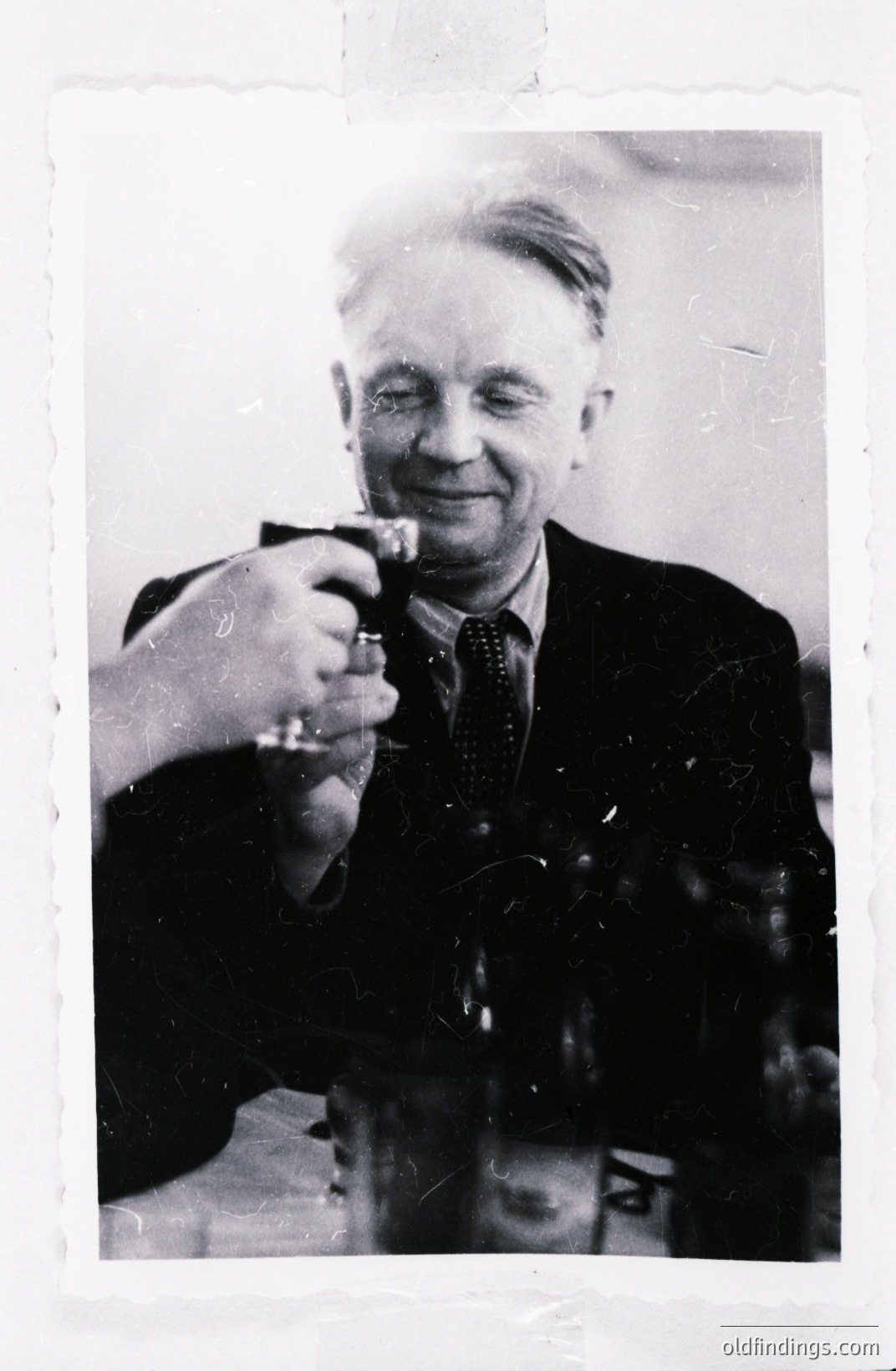 A monochrome image shows a smiling man raising a glass in a toast. He wears a dark suit and patterned tie, appearing to be in a restaurant or dining room setting. The table shows several bottles and glasses. Likely a candid, personal photograph.