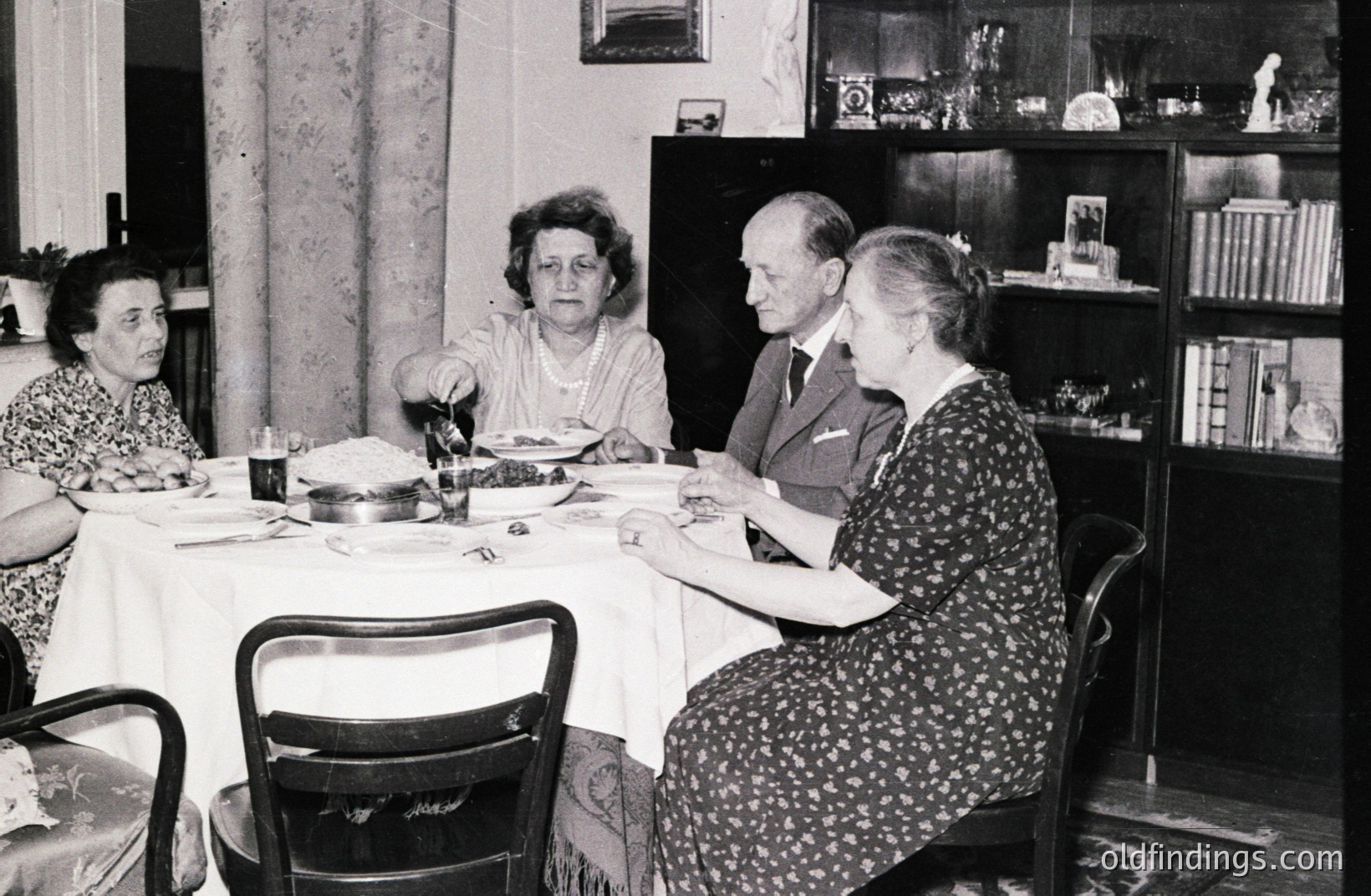 Family gathers around a table laden with food, likely a celebratory meal. The dining room features a built-in bookcase and patterned wallpaper. Likely a domestic scene, captured in black and white, possibly 1950s or 60s. Architectural details suggest a mid-century home. A typical family portrait.