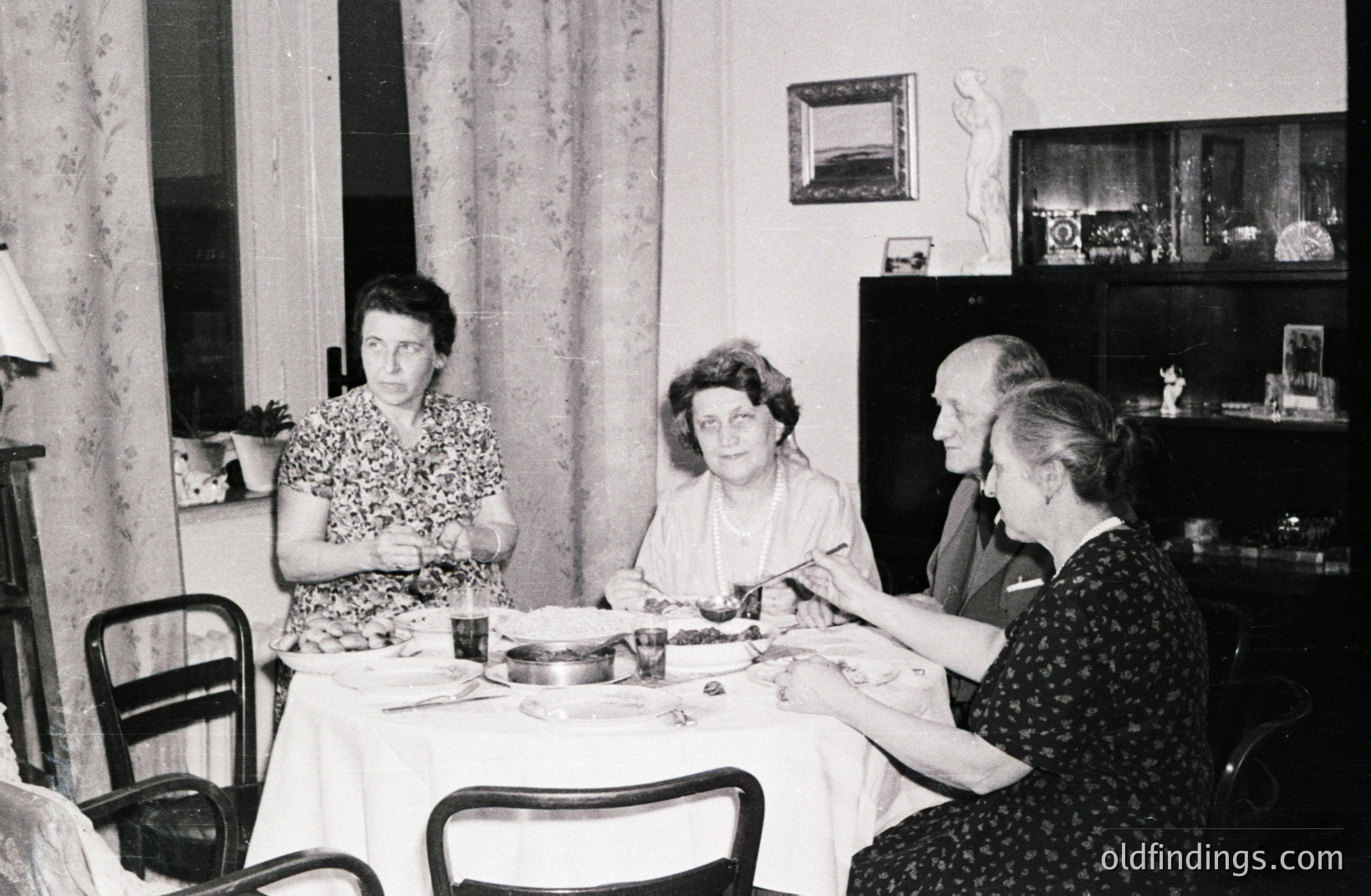 A formal dining scene: four individuals seated around a draped table, enjoying a meal. Notice the detailed cabinet decor and the classical bust statue. Likely a domestic interior from the 1950s or 1960s. Provides a window into mid-century family life.