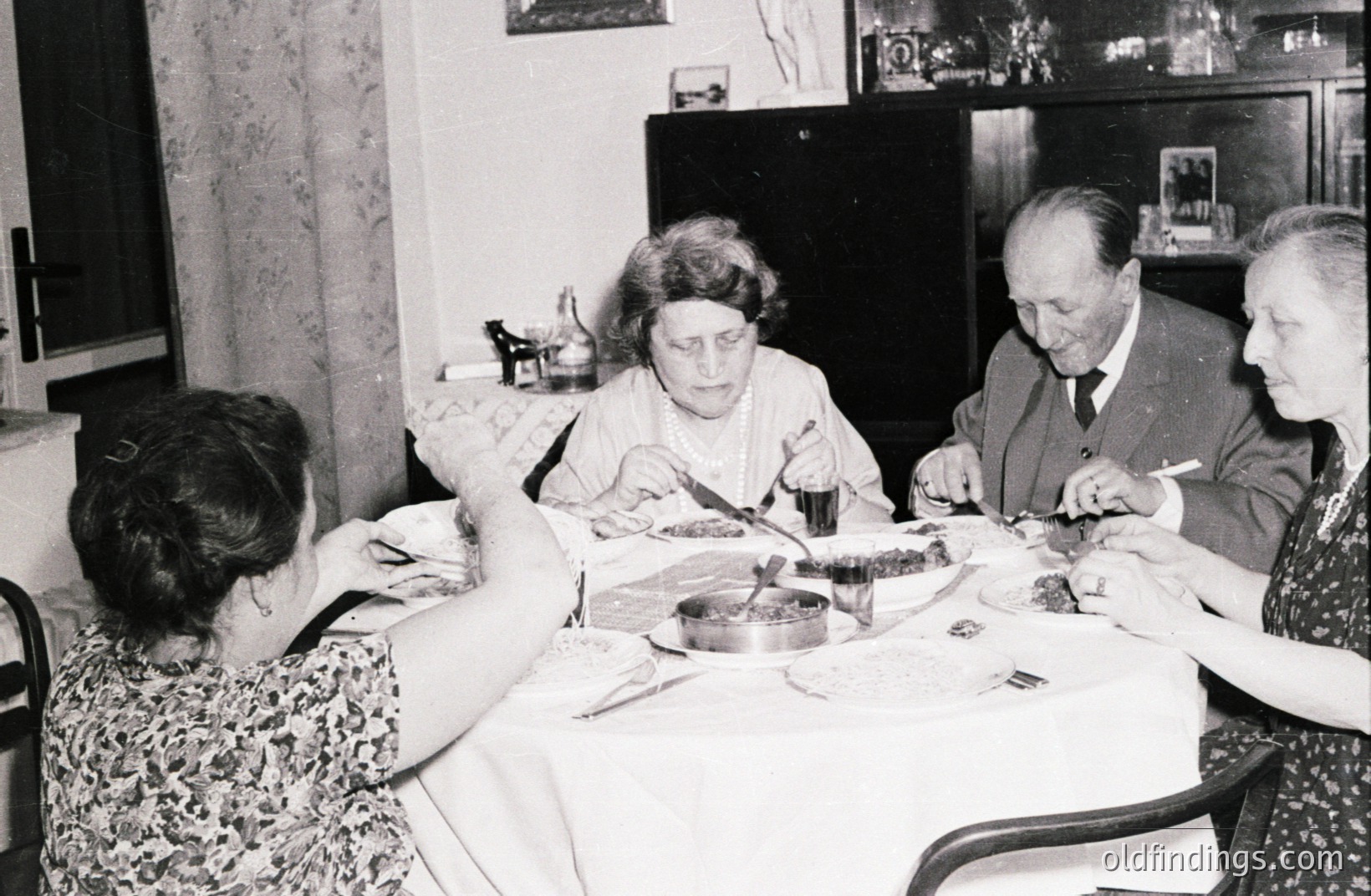 Four individuals seated around a table covered with a white tablecloth, appear to be enjoying a meal. The room features vintage decor including a credenza and framed artwork. Likely a family gathering, 1960s or 70s. Domestic scene with classic mid-century styling.