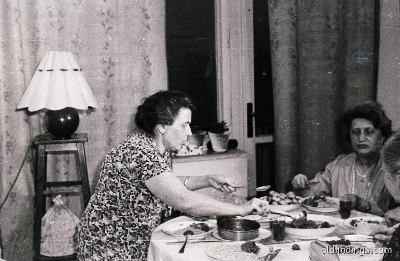 A family gathers for a formal indoor meal. A woman serves food from a pot with a spoon. Visible are plates, silverware, and a tablecloth-covered table. The setting features patterned wallpaper, a lamp, and drawn curtains. Likely a domestic scene, possibly 1950s-1960s.