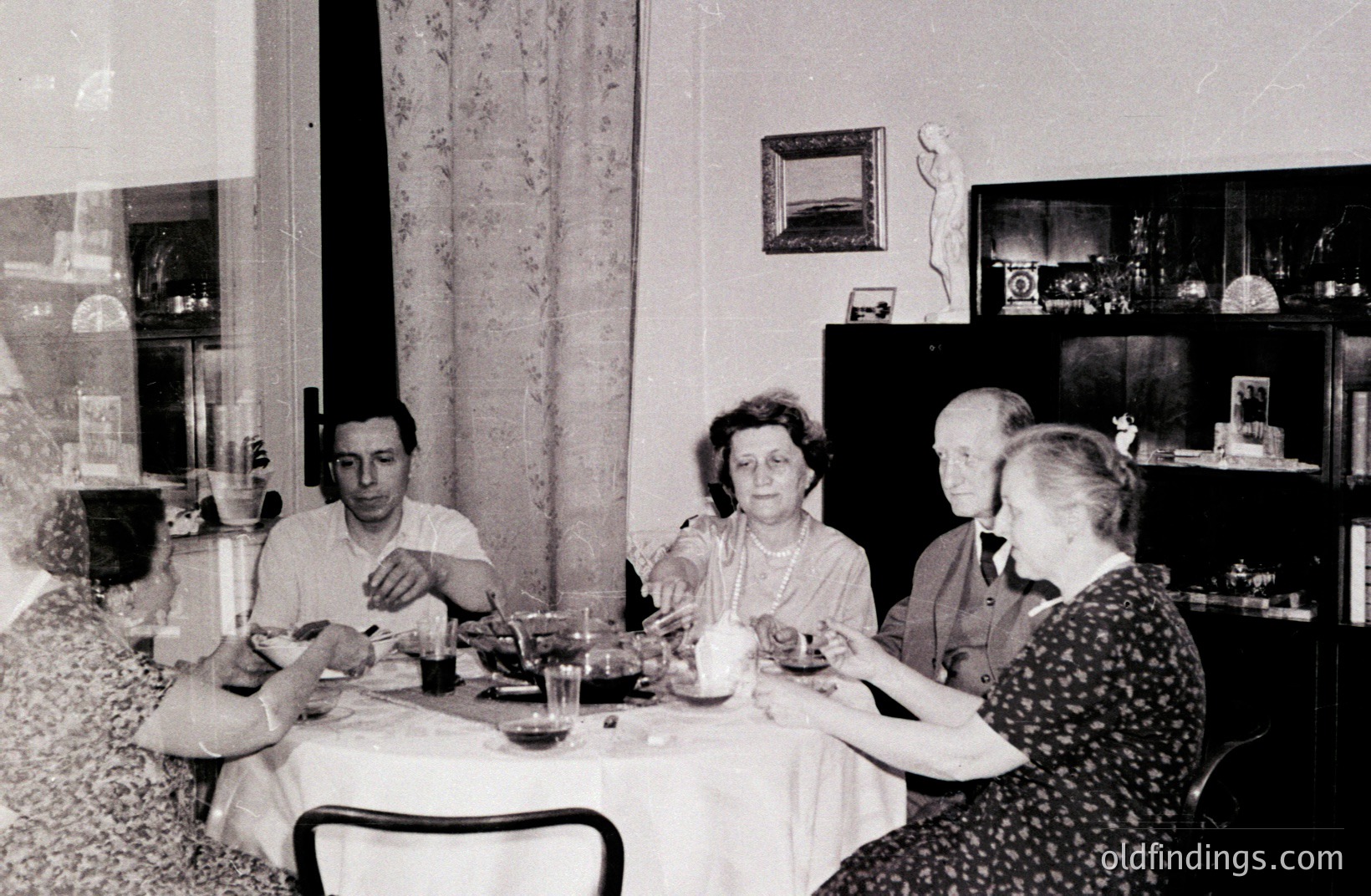 A seated group enjoys a domestic meal, possibly a tea or light lunch. The room features a sideboard, marble bust, and patterned curtains; suggesting a middle-class household. The photo likely dates to the 1940s-1960s, based on attire & photographic style.