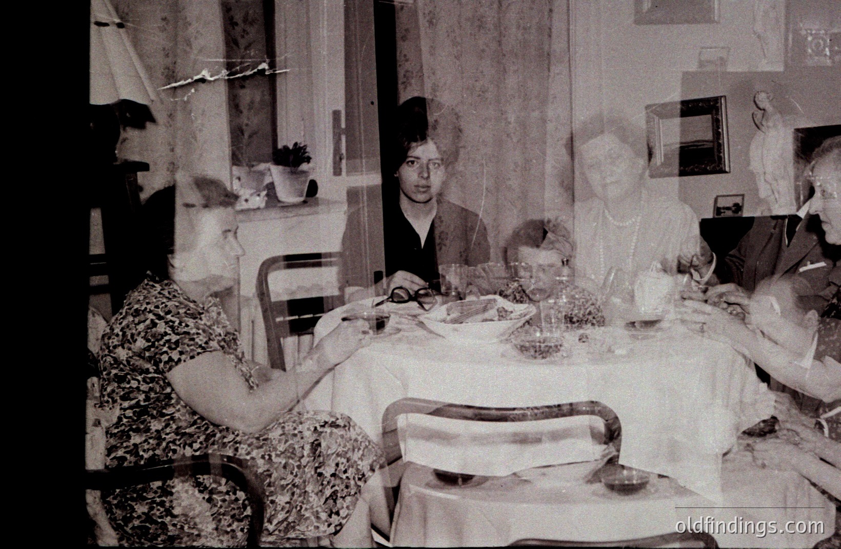A family gathers for a meal around a table covered in a patterned tablecloth. Visible are several generations, suggesting a multigenerational event. The décor indicates a home interior, possibly 1960s-70s. Subject matter valuable for family history or mid-century lifestyle studies.