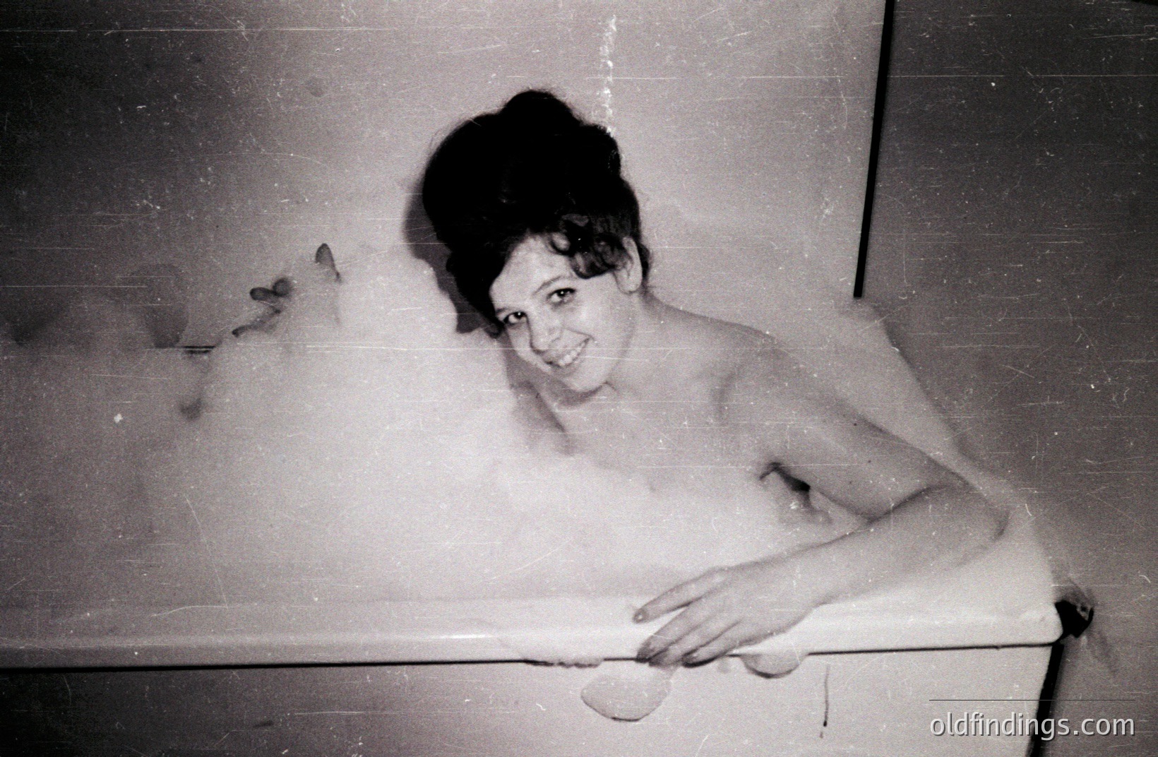 A woman with a stylized beehive hairstyle soaks in a frothy bathtub. She gazes directly at the camera with a slight smile. The photograph, likely a personal snapshot, displays signs of age with visible grain and scratches. Appears to be a 1960s domestic scene.