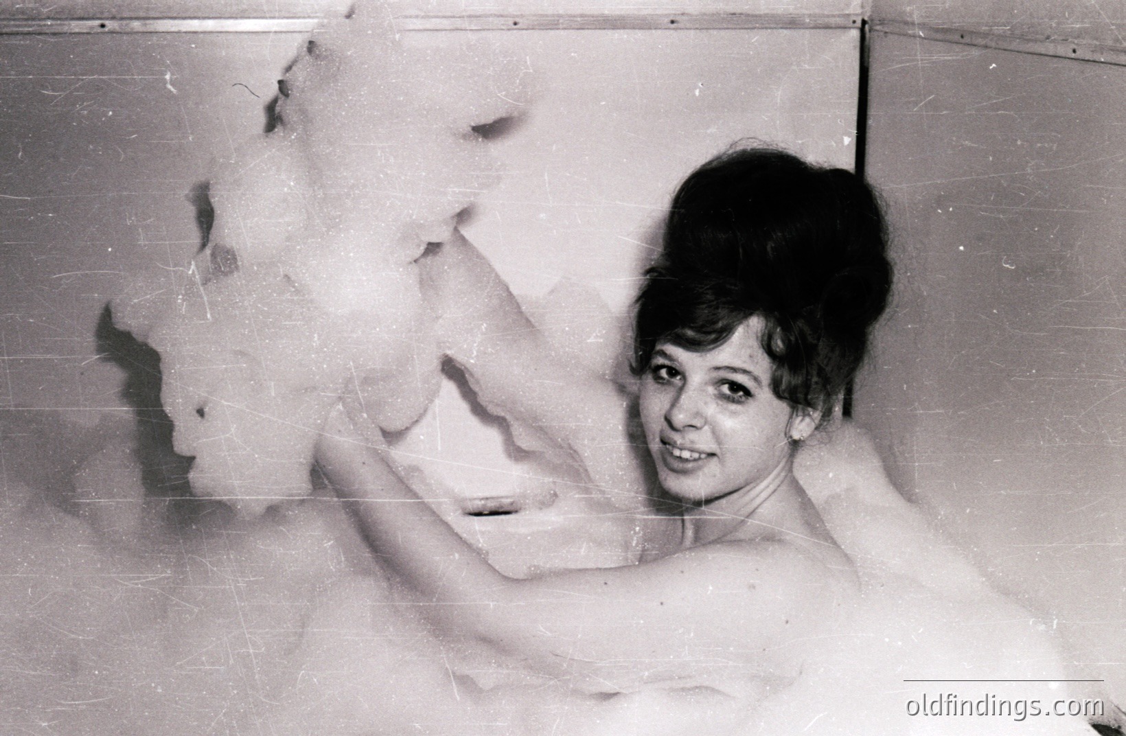 A woman with a beehive hairstyle is submerged in a large bubble bath, playfully gesturing toward the camera. The image, likely a studio portrait, showcases 1960s beauty standards and playful advertising. The composition is simple, emphasizing the bubble bath and the woman’s expression.