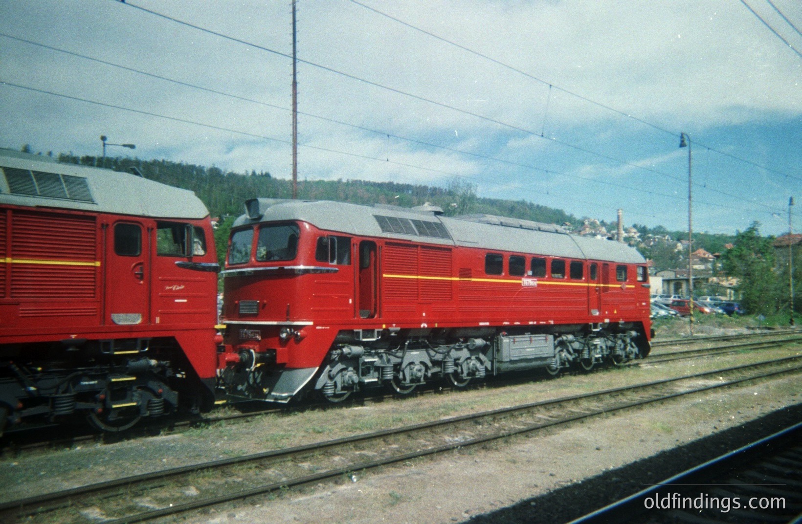 Two red diesel-electric locomotives sit on a railway line, likely in a depot or siding. The locomotives exhibit a boxy, streamlined design characteristic of mid-20th century European rail technology. Background features a hillside with vegetation and buildings. Possibly Bulgaria, 1960s-1970s. Ideal for transport design inspiration.