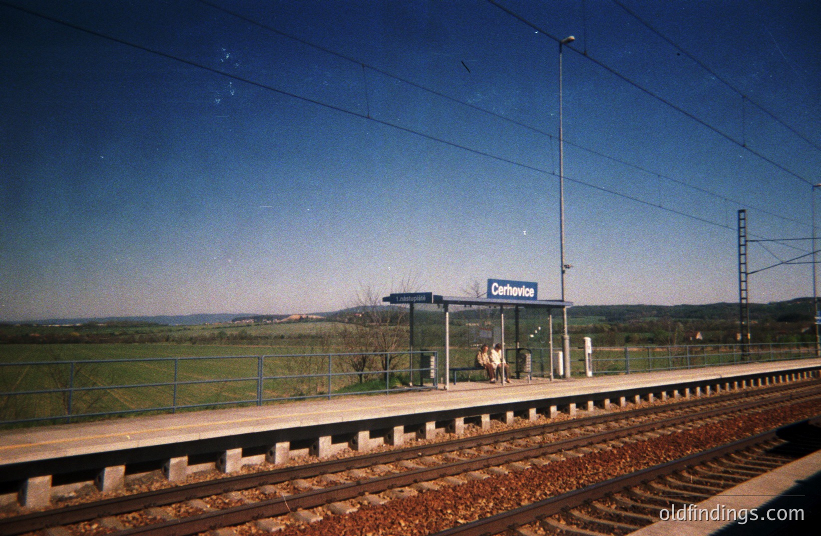 A solitary figure stands on the platform of the Černovice train station, framed by a simple, concrete shelter. Rolling hills and fields extend into the distance under a clear blue sky. Likely taken in the 1970s or 80s, showcasing a quiet, rural station scene. #Černovice