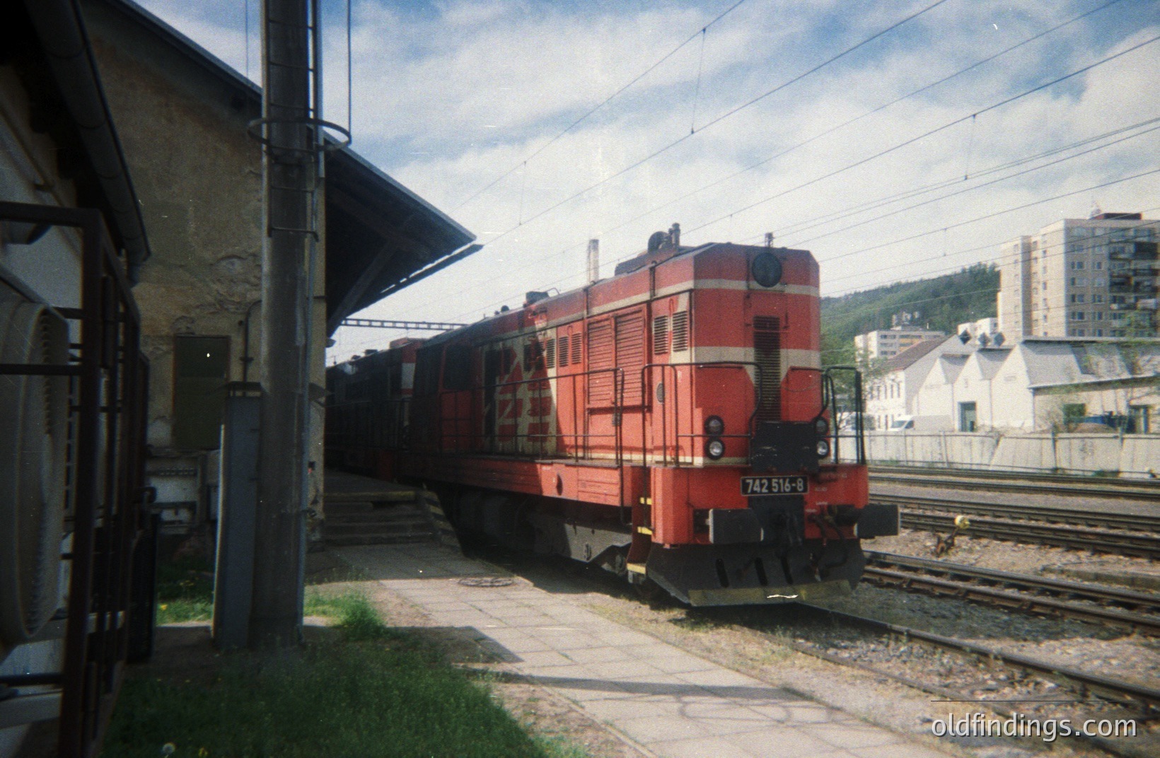 A red diesel locomotive (number 742 516-8) stands on tracks at a railway station platform, framed by a weathered station shelter. Background shows apartment buildings and green hillside. Likely Eastern European, potentially Bulgaria, based on locomotive style.