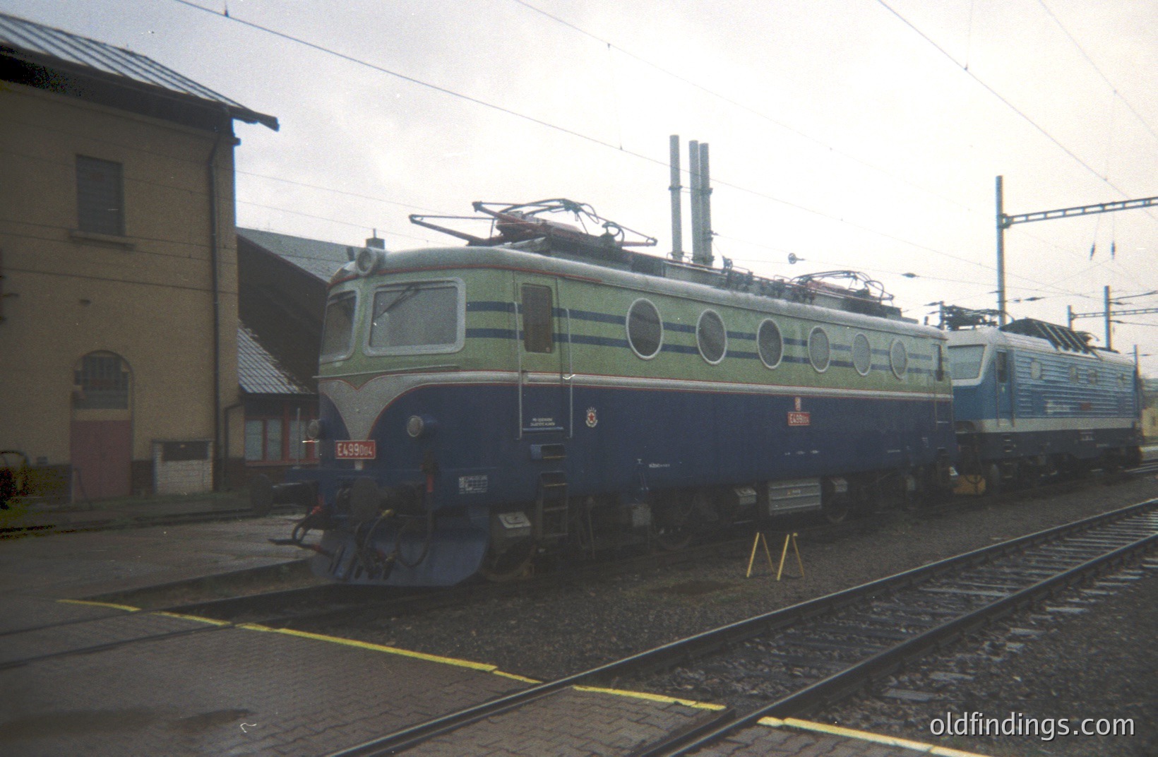 Striking image of a Swiss Federal Railways (SBB) electric locomotive, likely an Ae 4/7 series, detailed with green and blue livery & circular ventilation ports. Station building visible in the background. Classic mid-century railway infrastructure.