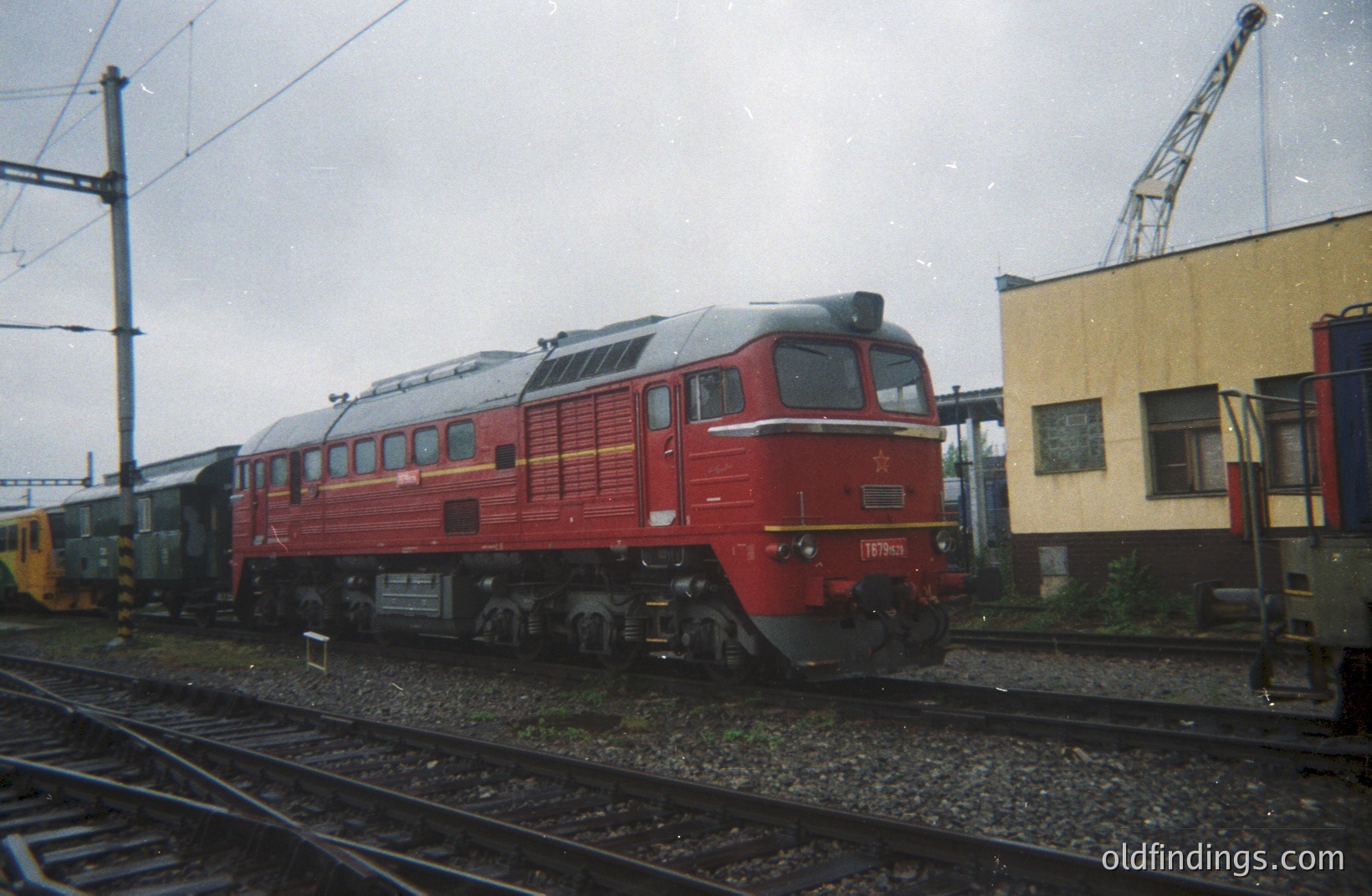A red, Soviet-era diesel locomotive (likely a TE1 series) sits on tracks within a railway depot. The number 17630 is visible on the front. Freight cars and a section of a yellow passenger carriage are partially visible. A crane is visible near a building.