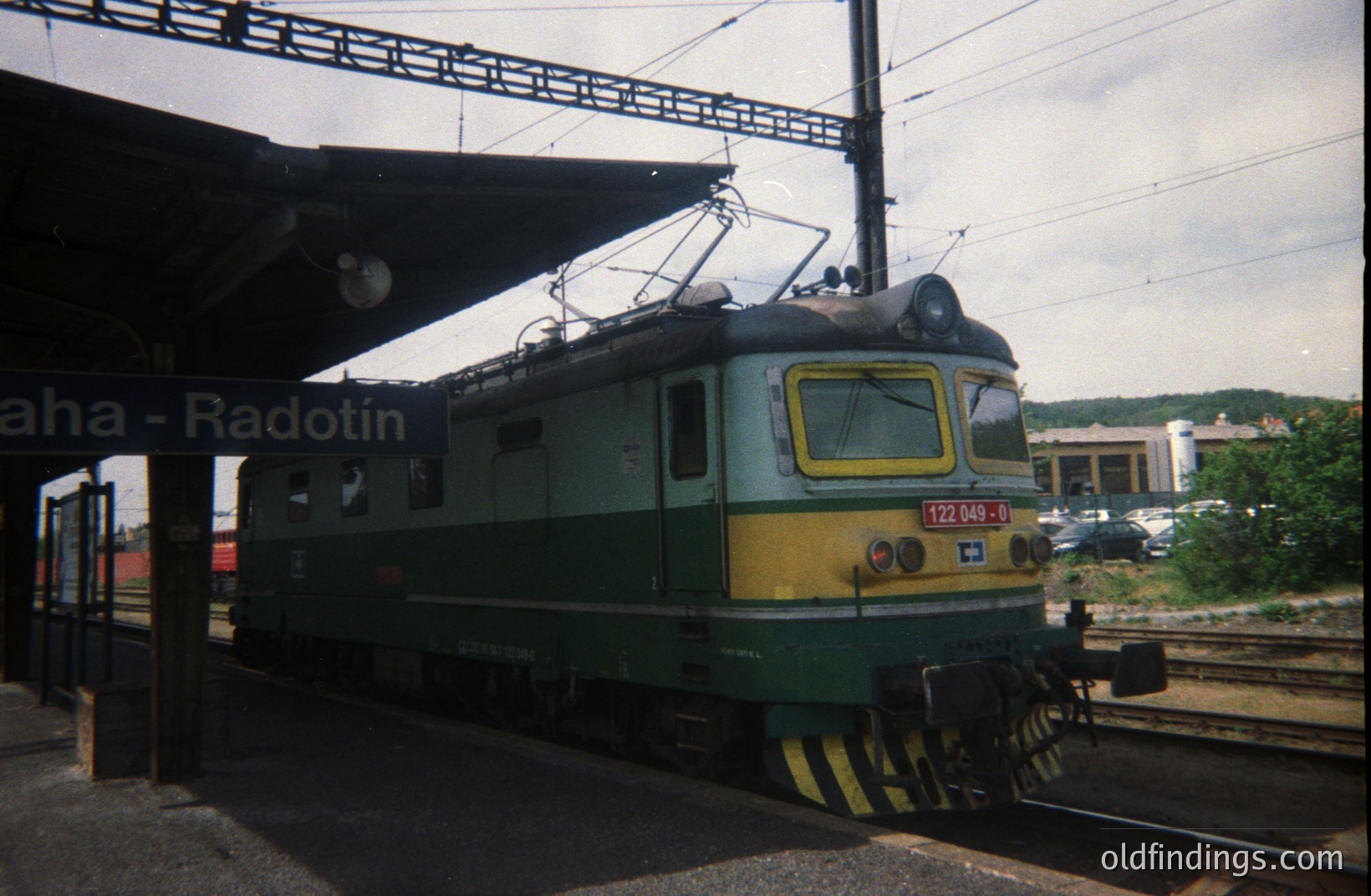 Bulgarian State Railways electric locomotive (type 122, number 049) at Radotin station. Likely captured in the 1970s-80s given the era-appropriate locomotive and color scheme. Platform overhead protection visible. A rural landscape is visible in the background.