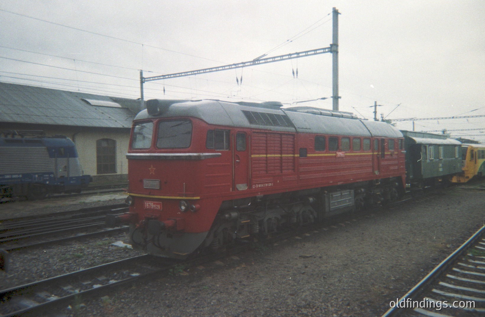 A red diesel locomotive, likely a Soviet-era M62 model, sits on tracks alongside a green passenger car. The scene is industrial, with a station building visible in the background. Possible location: Eastern Europe. Likely timeframe: 1970s-1980s. Excellent for rail history or transportation design.