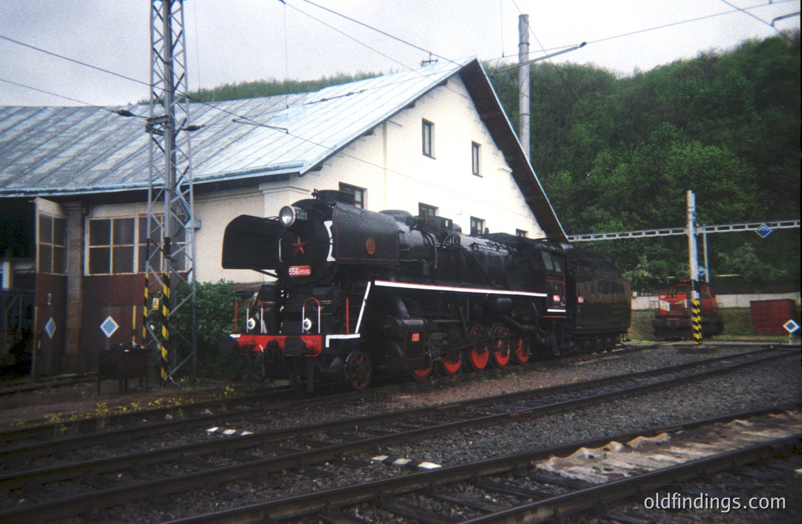 A stationary, black steam locomotive rests on tracks beside a functional rail yard building with a metal roof and visible electrical infrastructure. Lush green foliage fills the background. The locomotive's markings are partially obscured, possibly indicating a former industrial use. Likely taken in the 1970s or 80s.