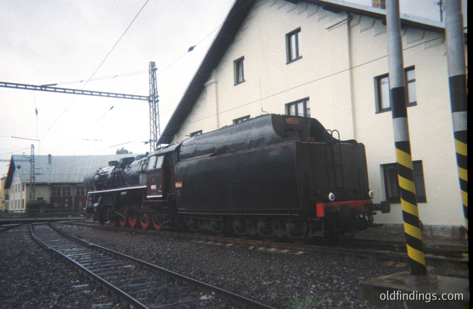 A weathered, stationary steam locomotive rests on tracks beside a plain, industrial building. The locomotive exhibits dark green and black coloring with visible wear. Likely 1970s-80s, judging by the film stock and industrial setting. A safety pole stands prominently in the foreground.