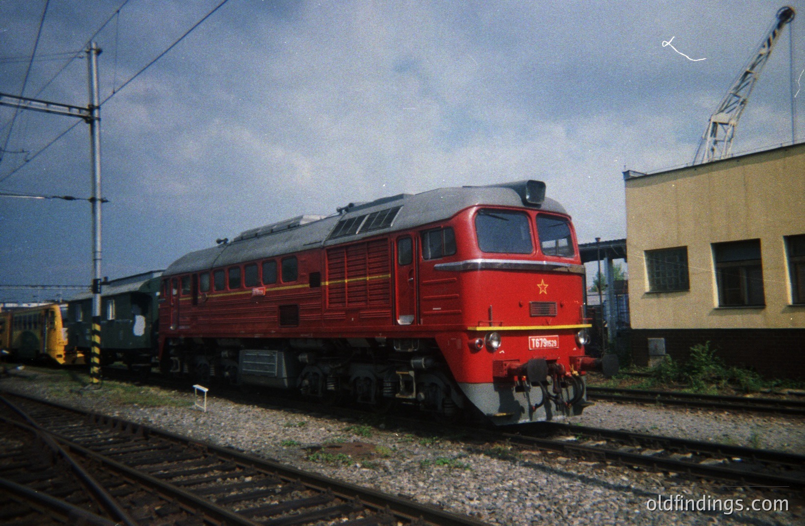 A powerful, red Soviet-era diesel locomotive (likely a TE3 series) sits on tracks, coupled to freight cars. The locomotive number 197903 is visible. A rail yard setting with a control tower visible in the background. Appears to be mid-1970s.