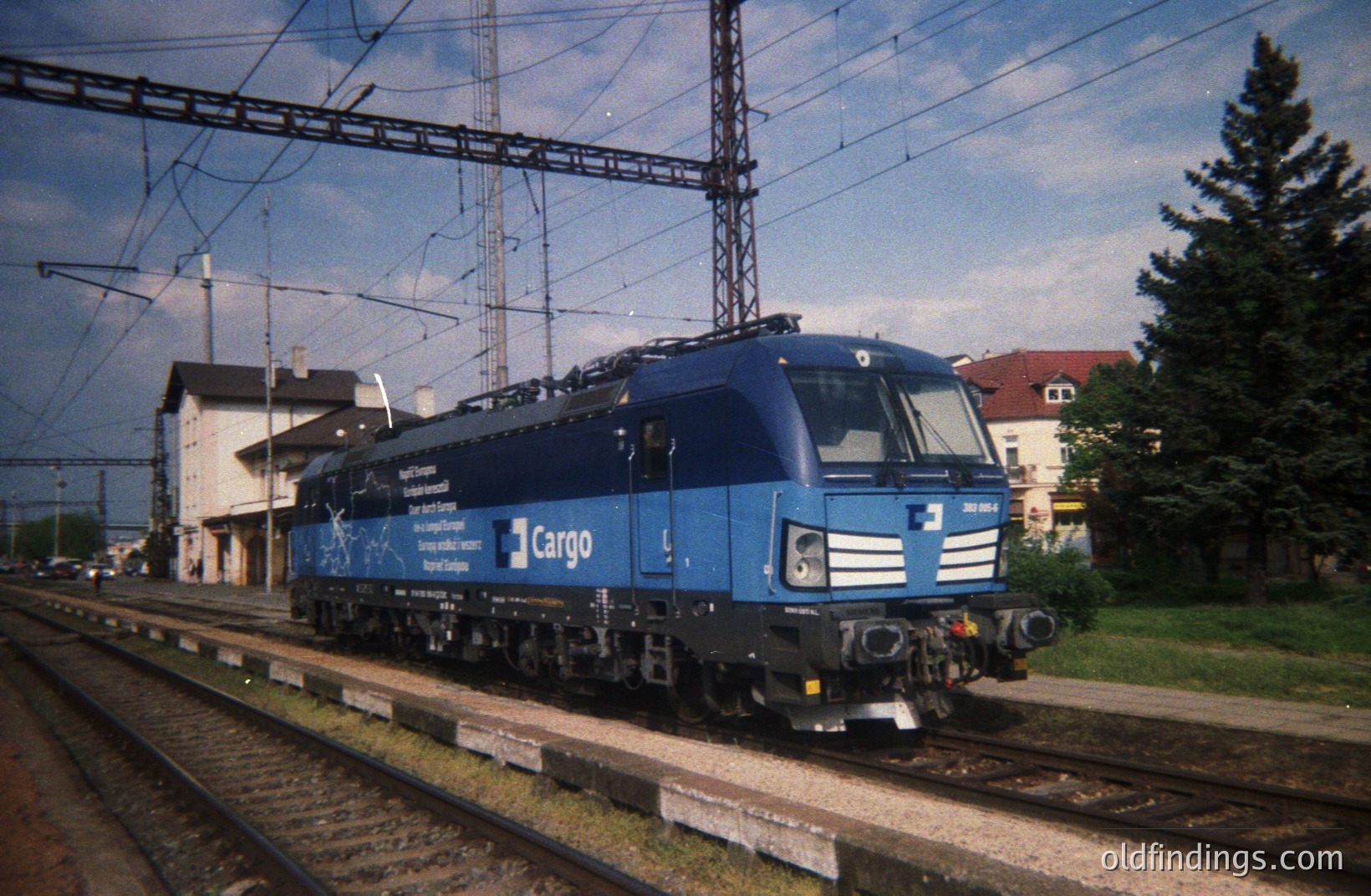 A blue electric locomotive, likely a Siemens ES64U2, stands on a track in a rail yard. The locomotive displays "CargoNet" branding. A two-story brick building with a gabled roof sits in the background. Appears to be a Central European location. Mid-1990s - early 2000s timeframe. Potentially useful for rail transport design.
