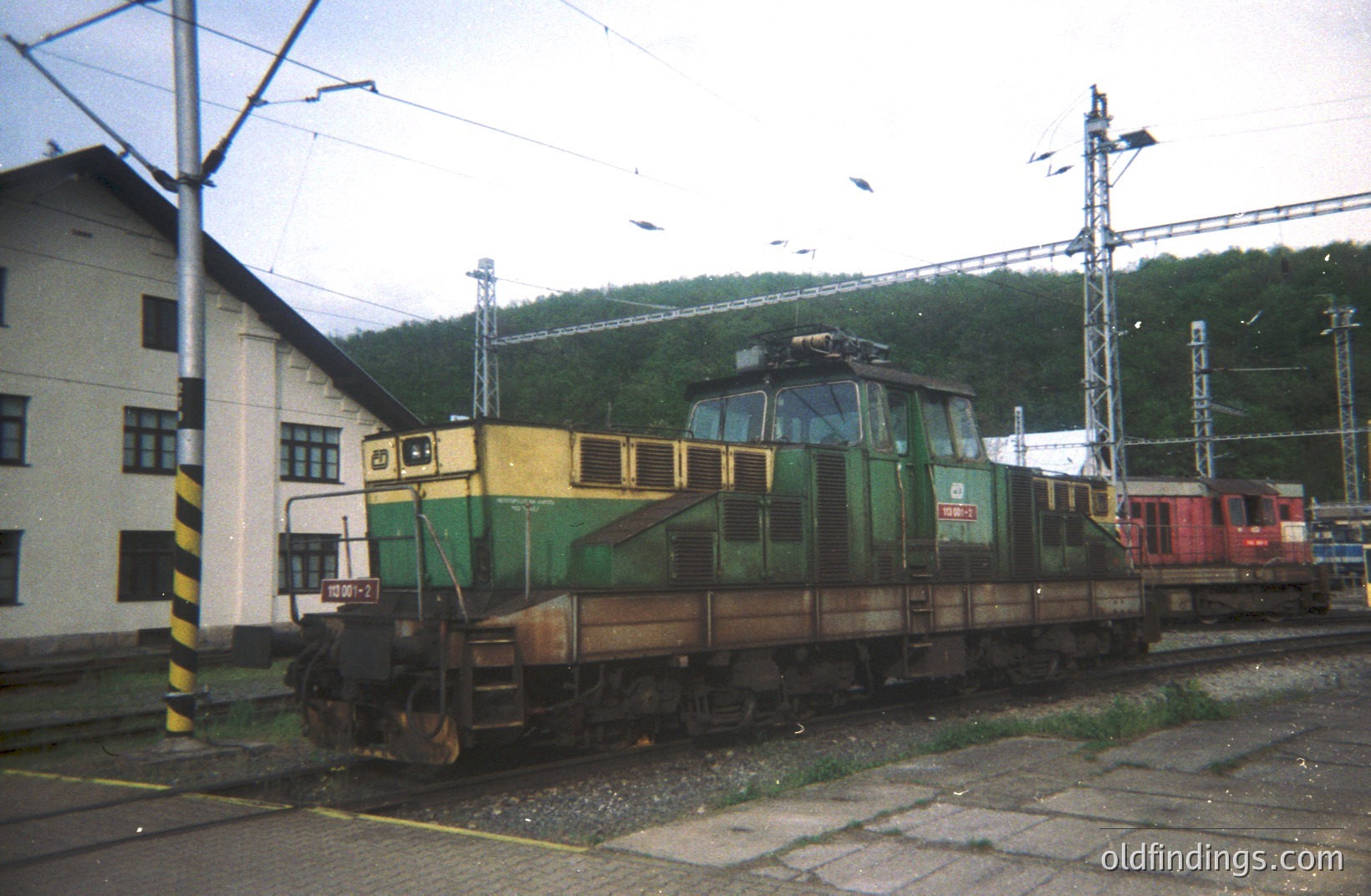 An overhead view depicts a Swiss Electric Locomotive (SBB Re 4/4) series, numbered 10602. It's situated on a rail yard next to a building. The locomotive features a yellow nose and green body. Background shows hillside vegetation and railway infrastructure. Likely 1960s-1970s, based on the train’s design. A valuable resource for railway history.