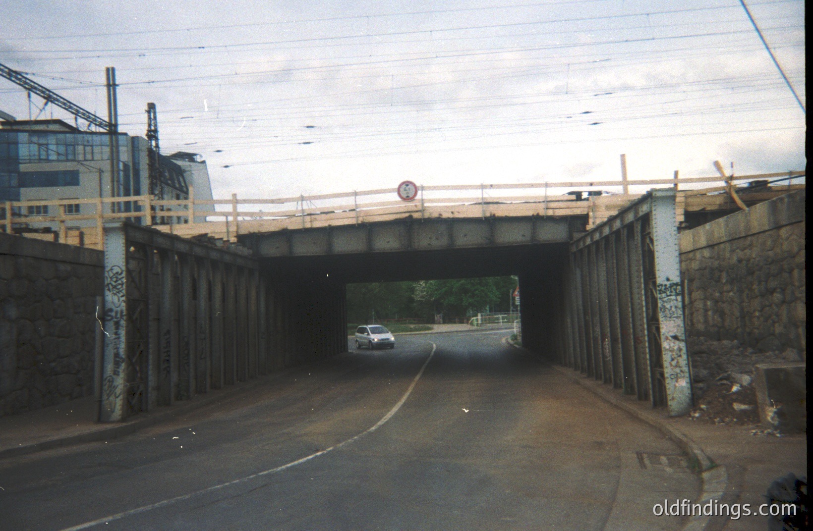 Overpass view: a single car traverses a roadway tunnel lined with weathered stone pillars & graffiti. A modern building rises in the background, contrasted against a cloudy sky. Likely a post-industrial landscape, potential for urban studies or architectural reference.