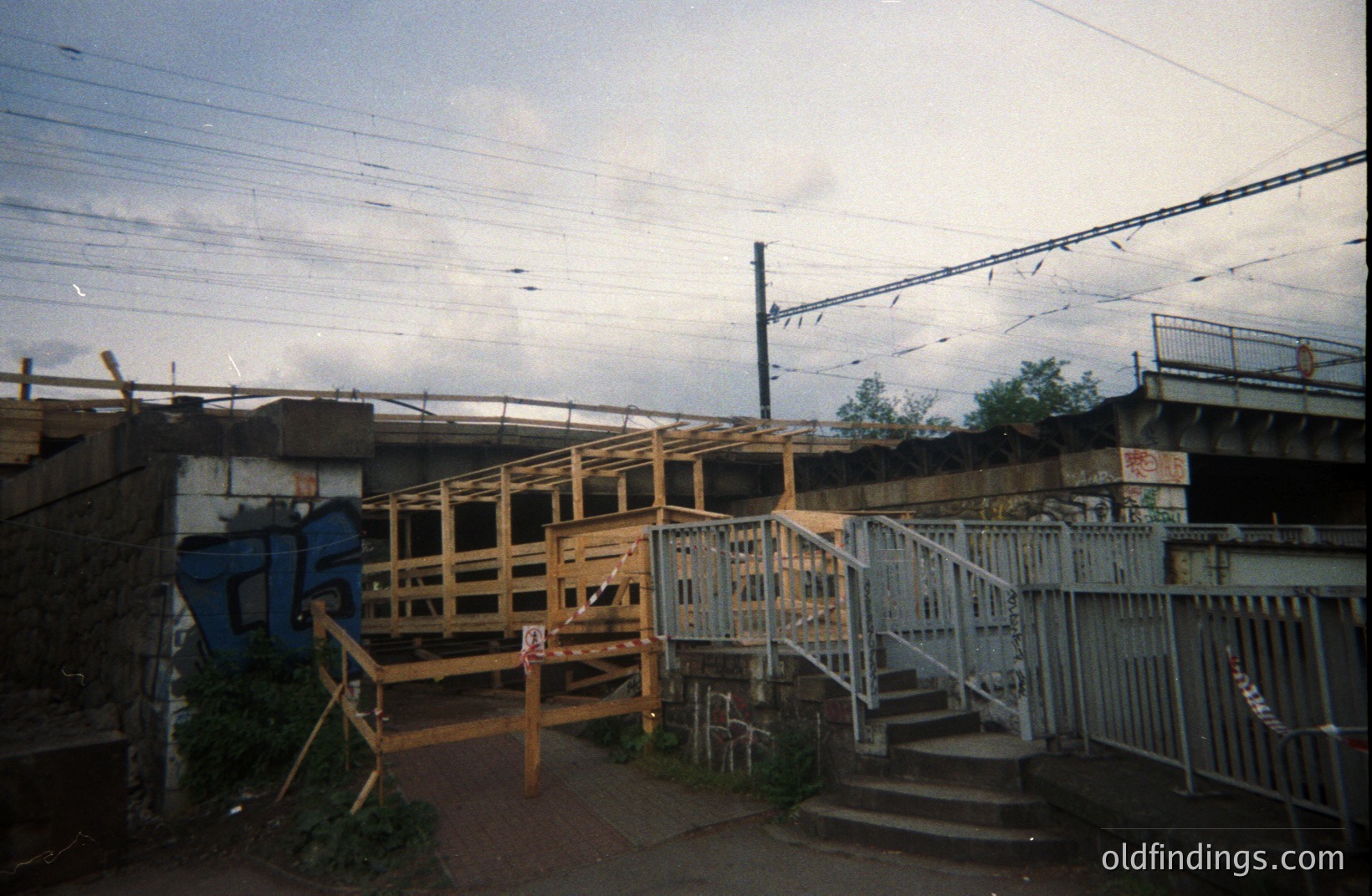 Overpass under construction, likely a pedestrian bridge, framed with exposed lumber & scaffolding. Railway lines and embankment visible overhead. Brickwork displays faded graffiti. Appears to be a working-class urban area. Likely 1980s-1990s.