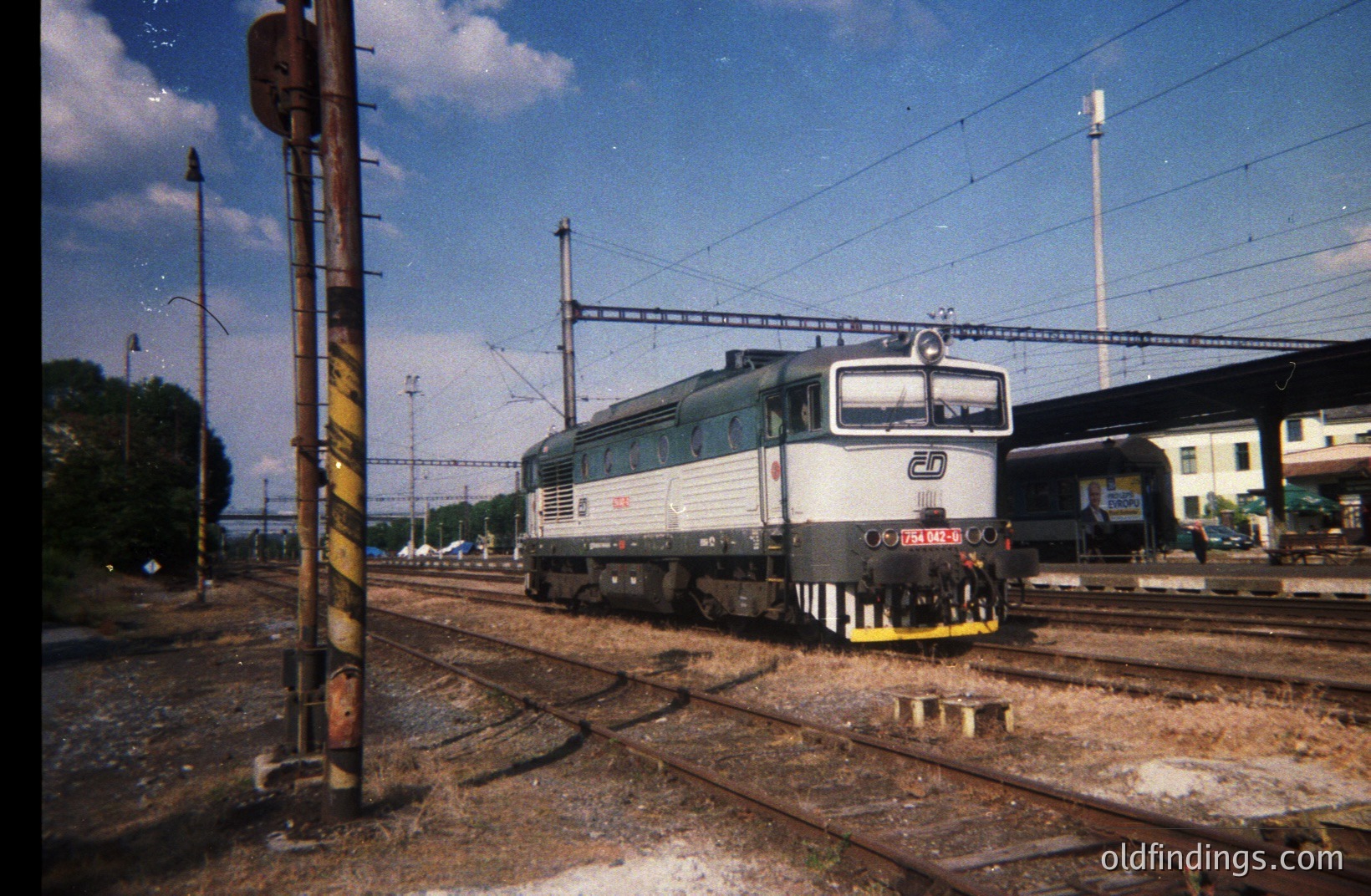 Detailed view of a green diesel locomotive, possibly a class 66, on a rail line. Background shows a station platform, greenery, and sky. Train number visible as "66 174 603". Likely photographed in Europe, 1990s timeframe. Rail infrastructure visible.