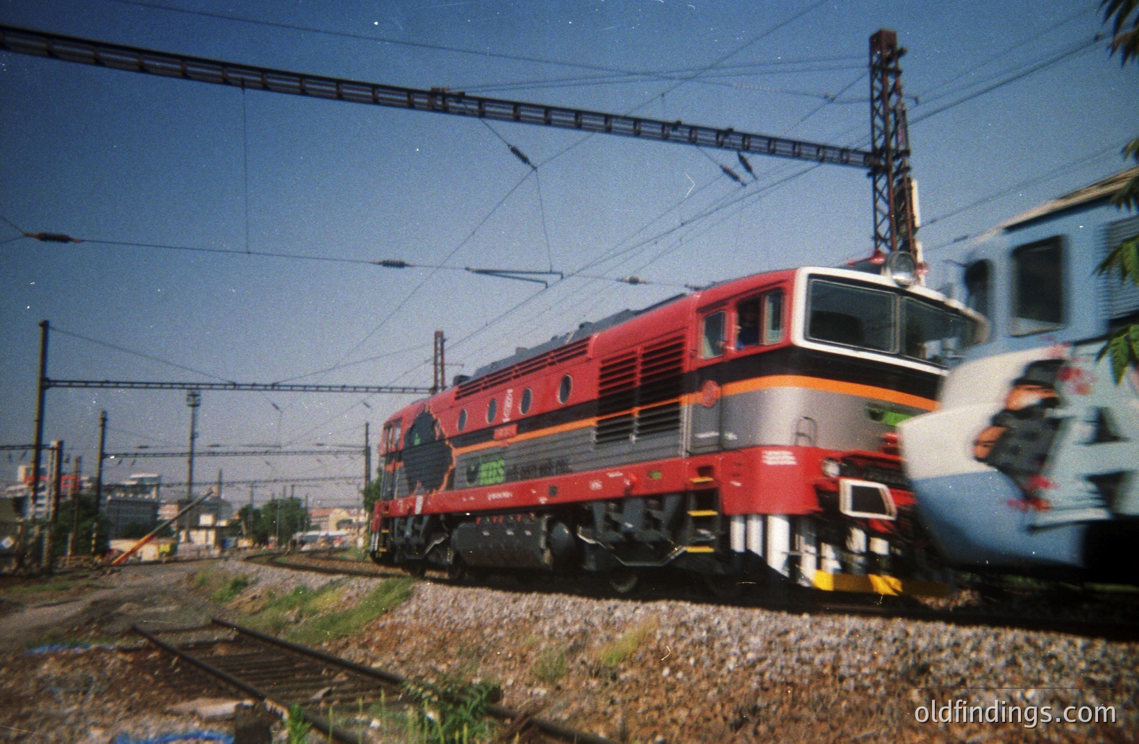 A red and grey electric locomotive, likely a BD series, moves along a track with overhead lines, typical of Bulgarian railways. Graffiti covers a railcar in the background. Gravel and sparse vegetation line the rails. Likely taken in the 1980s or 1990s.