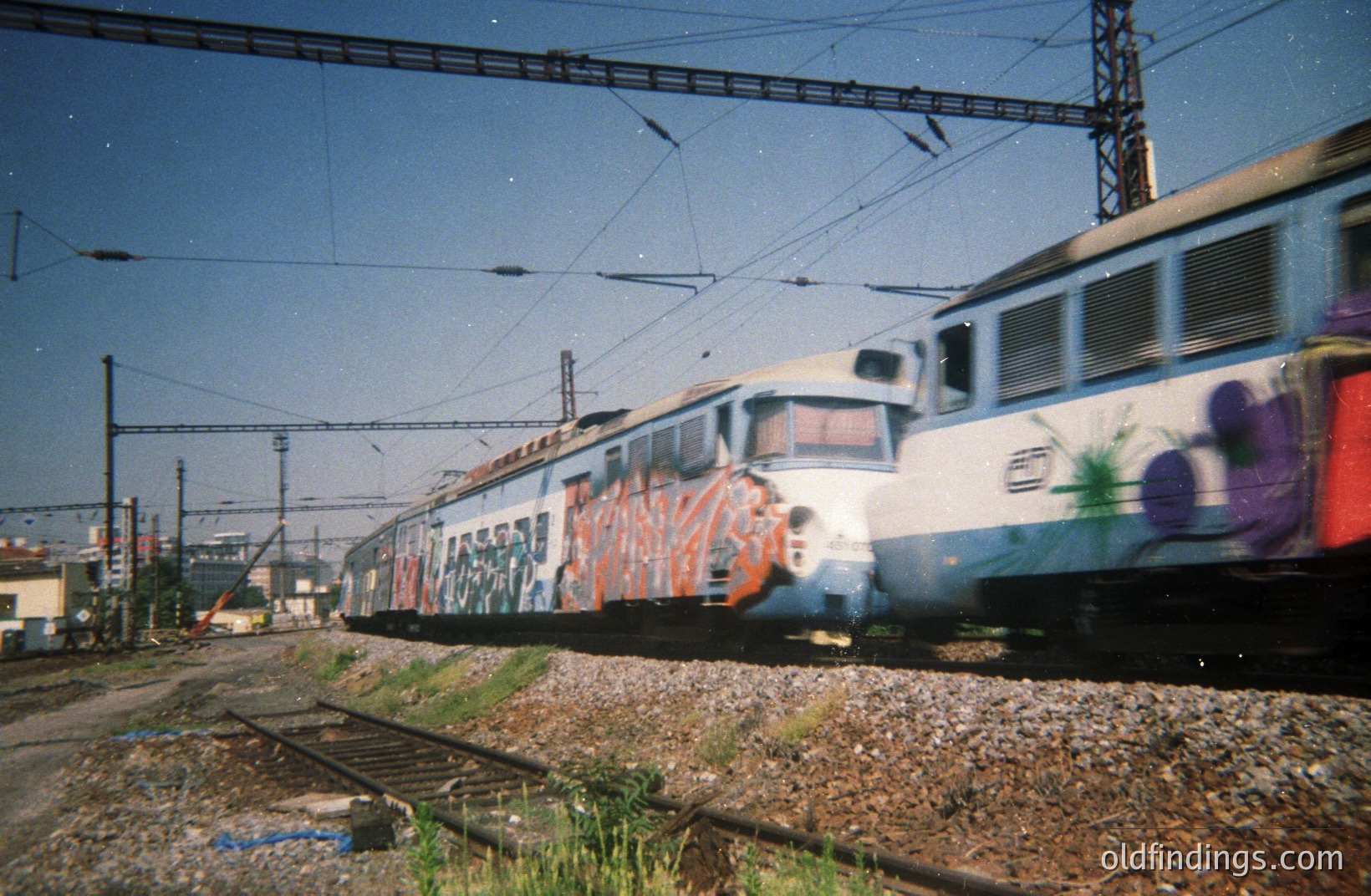 A high-speed passenger train, likely electric, speeds along tracks with vibrant graffiti covering its exterior. The scene suggests a rail yard or industrial area. Appears to be a candid, likely 1980s or 90s snapshot capturing urban art and transportation. A striking visual document.