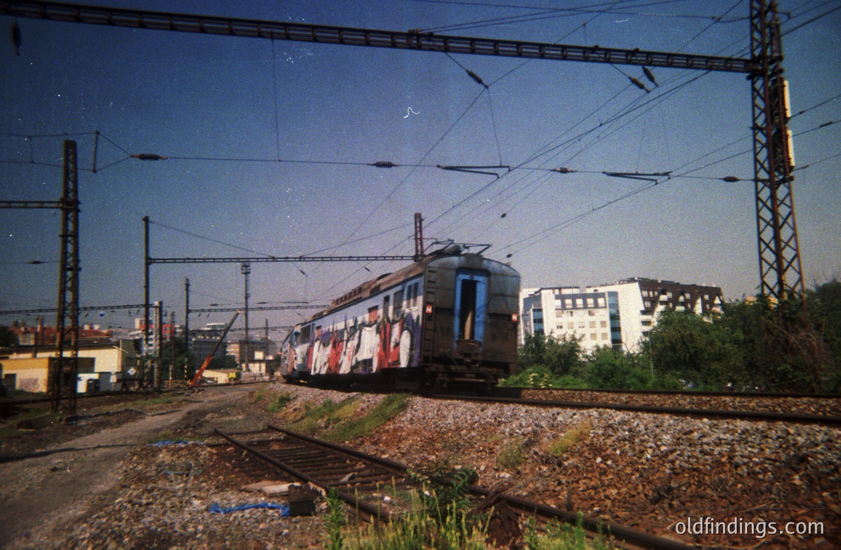 A graffiti-covered electric train passes along a track, framed by overhead wires and utility poles. Visible are residential buildings, suggesting an urban or suburban location. The photo’s film grain and color palette imply a 1980s or 1990s timeframe. Could be used for urban design references.
