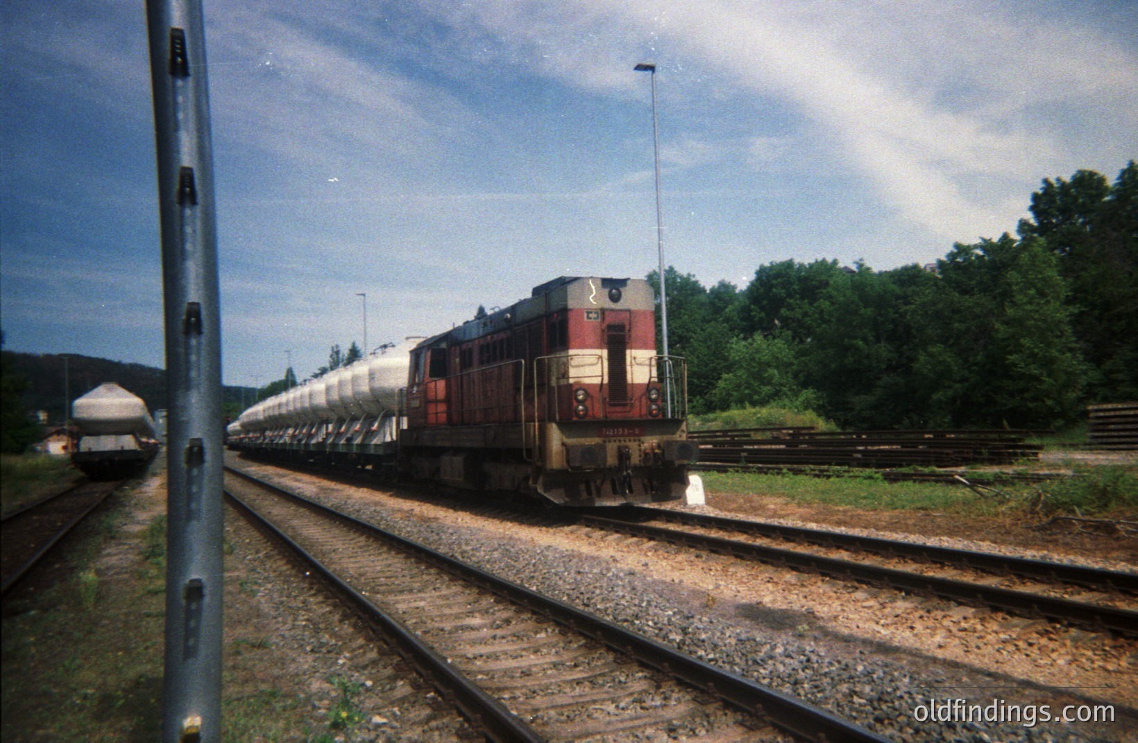 Long freight train with a red and gray locomotive, likely a diesel, sits on a gravel track bed alongside a grassy embankment. The train features several tank cars. A utility pole dominates the left foreground. Lush greenery fills the background. Appears to be a rural industrial area. Likely 1970s-1980s.