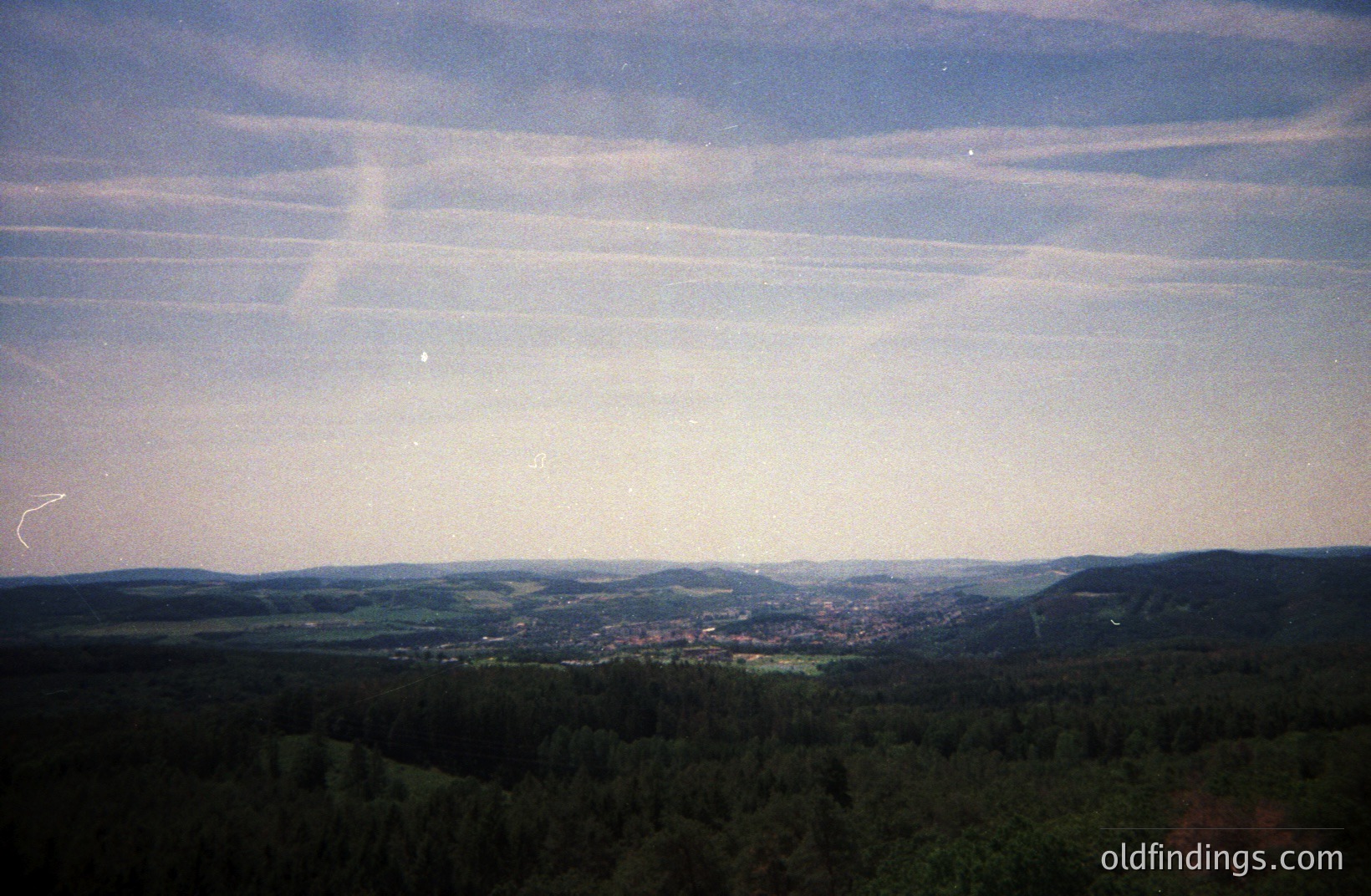 Elevated view of a valley town nestled amongst forested hills. Prominent contrail patterns dominate the sky, hinting at aerial activity. Appears to be a late afternoon or early evening scene with soft, diffused light. Possible European location.