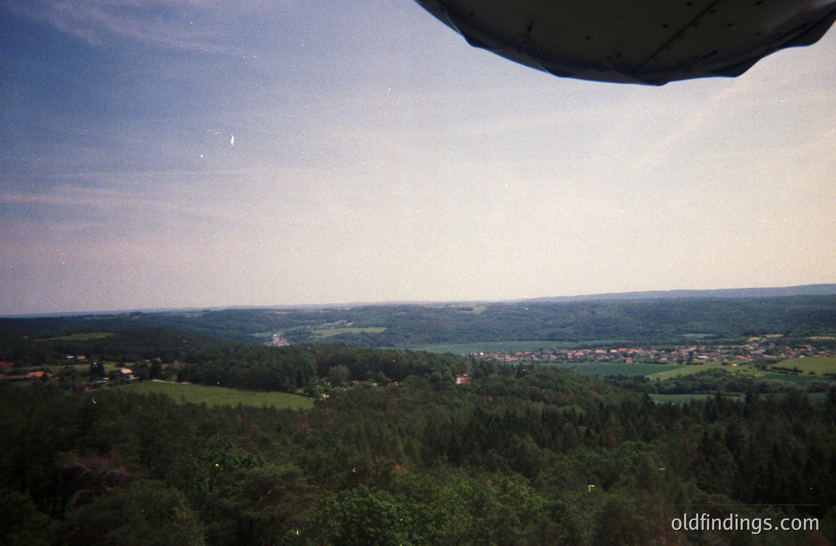 Aerial view from a plane window showcases a forested landscape transitioning to a town in the distance. The scene features a broad, green valley and rolling hills beneath a partly cloudy sky. Appears to be a rural European setting. Likely 1990s or early 2000s based on film aesthetic.