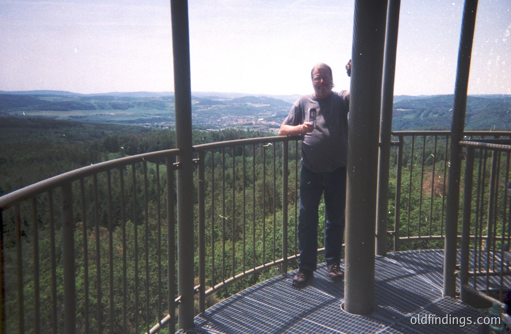 A man stands on a circular metal observation platform, overlooking a sprawling forested valley and distant town. He wears a dark t-shirt and jeans, appearing relaxed with hands on his hips. The platform’s metal grates and railing are visible. Likely a tourist snapshot.